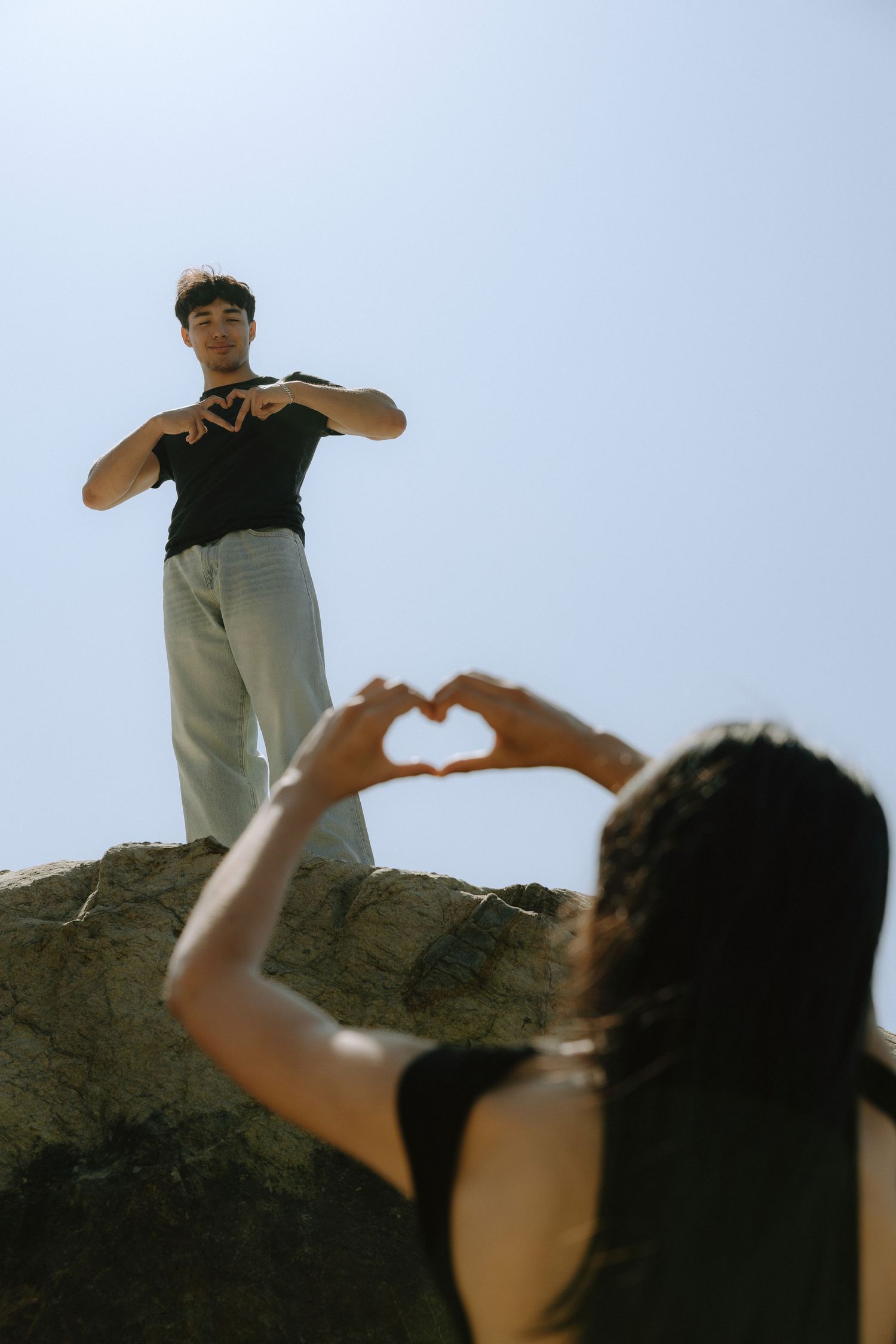 Two people making heart shapes with their hands, one standing on a rock against a clear blue sky.