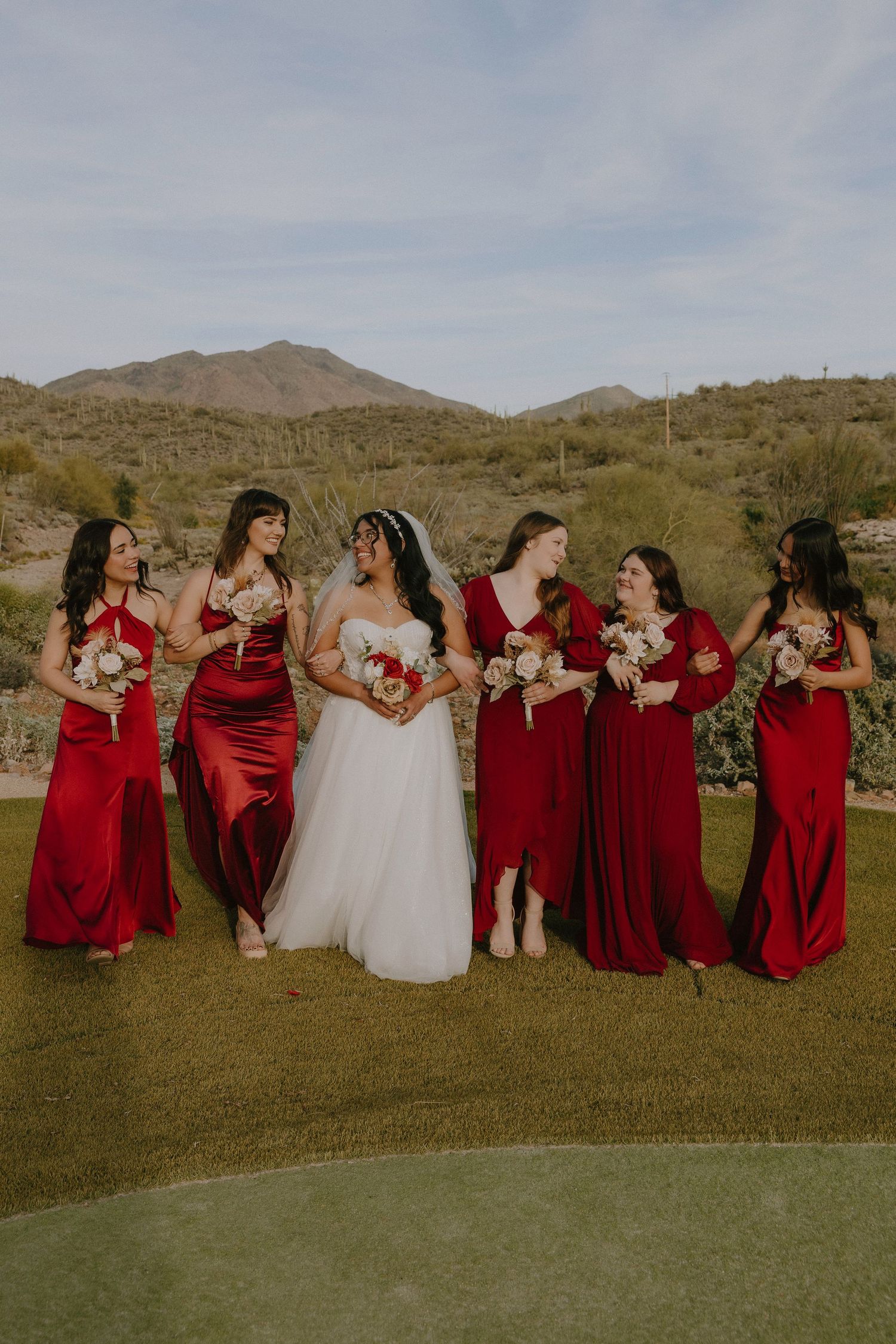 Bride in white gown with five bridesmaids in red satin dresses holding bouquets, posing outdoors with desert mountain backdrop.