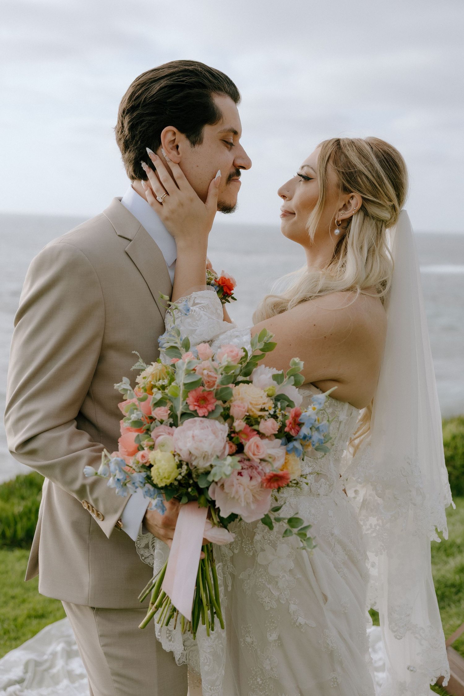 Bride holding colorful bouquet gazes at groom in tan suit with ocean backdrop at outdoor wedding.