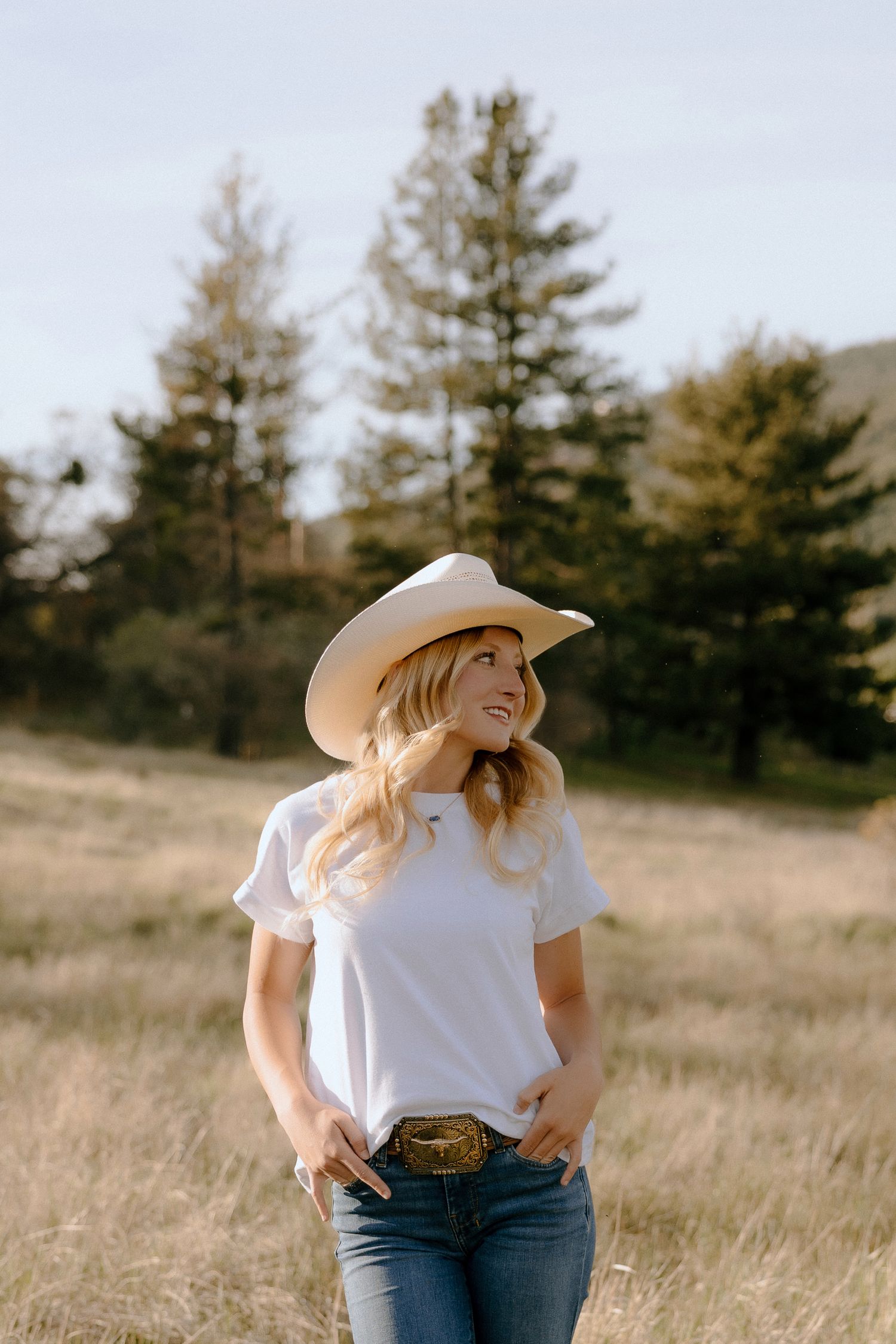 Smiling blonde woman in white tee, cowboy hat, and jeans stands in a sunlit field with pine trees behind her.