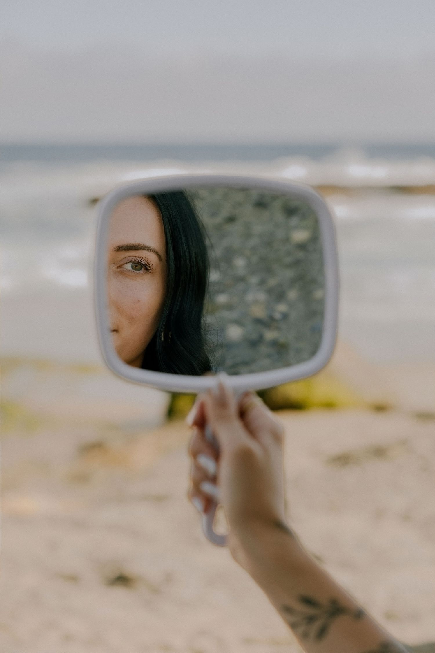 A hand with a tattoo holds a small mirror reflecting a young woman's face on a sandy beach.