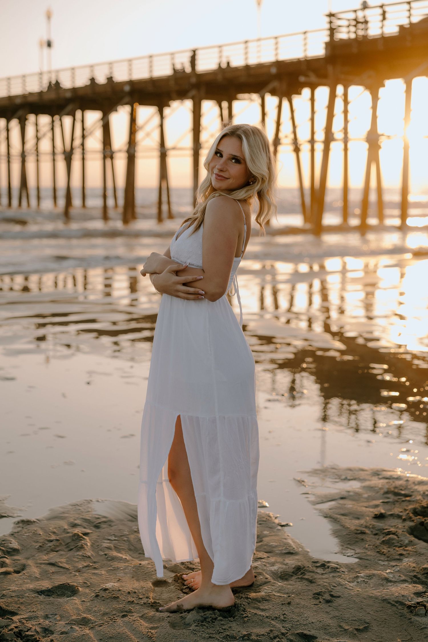 Blonde woman in white maxi dress poses barefoot on beach at golden sunset near a wooden pier.
