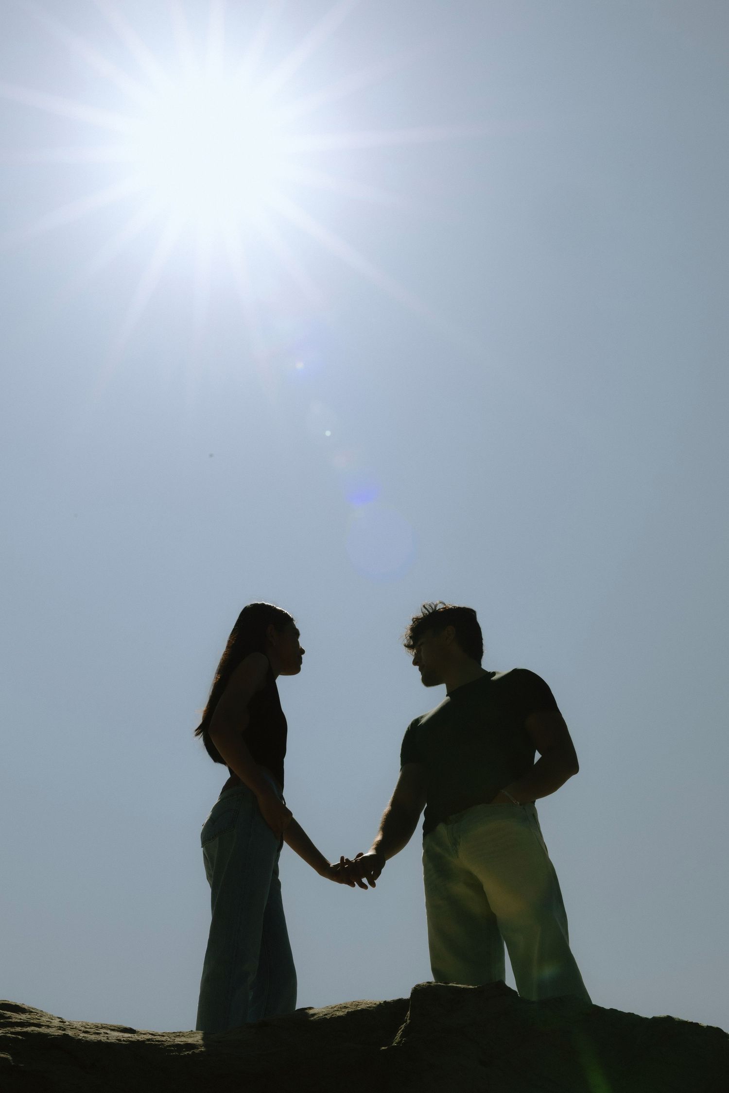 Romantic silhouette of couple holding hands on rocks against bright sunlit sky with lens flare effect.