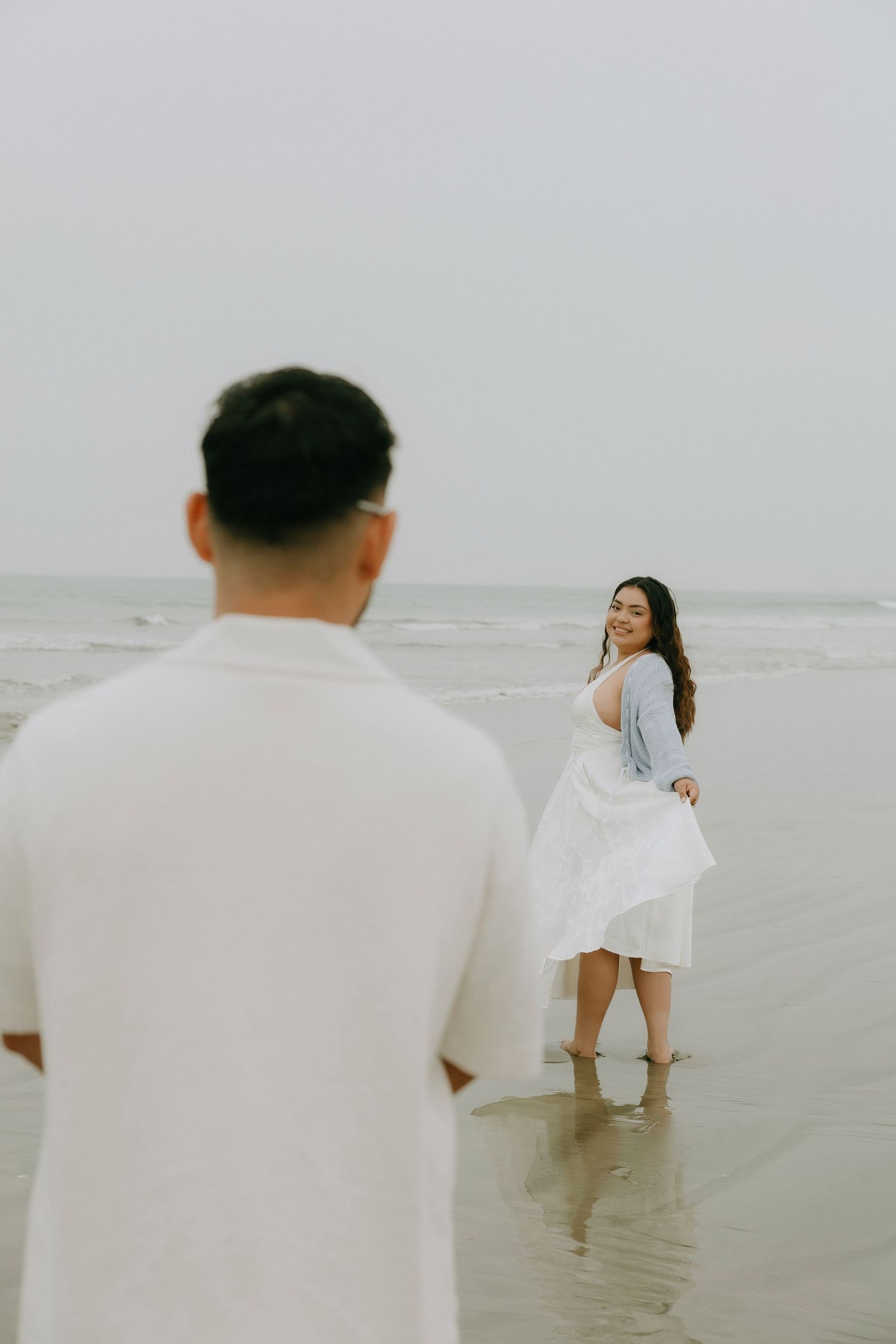 Couple on a beach, woman in white dress smiling back at man in white shirt, ocean waves in background.