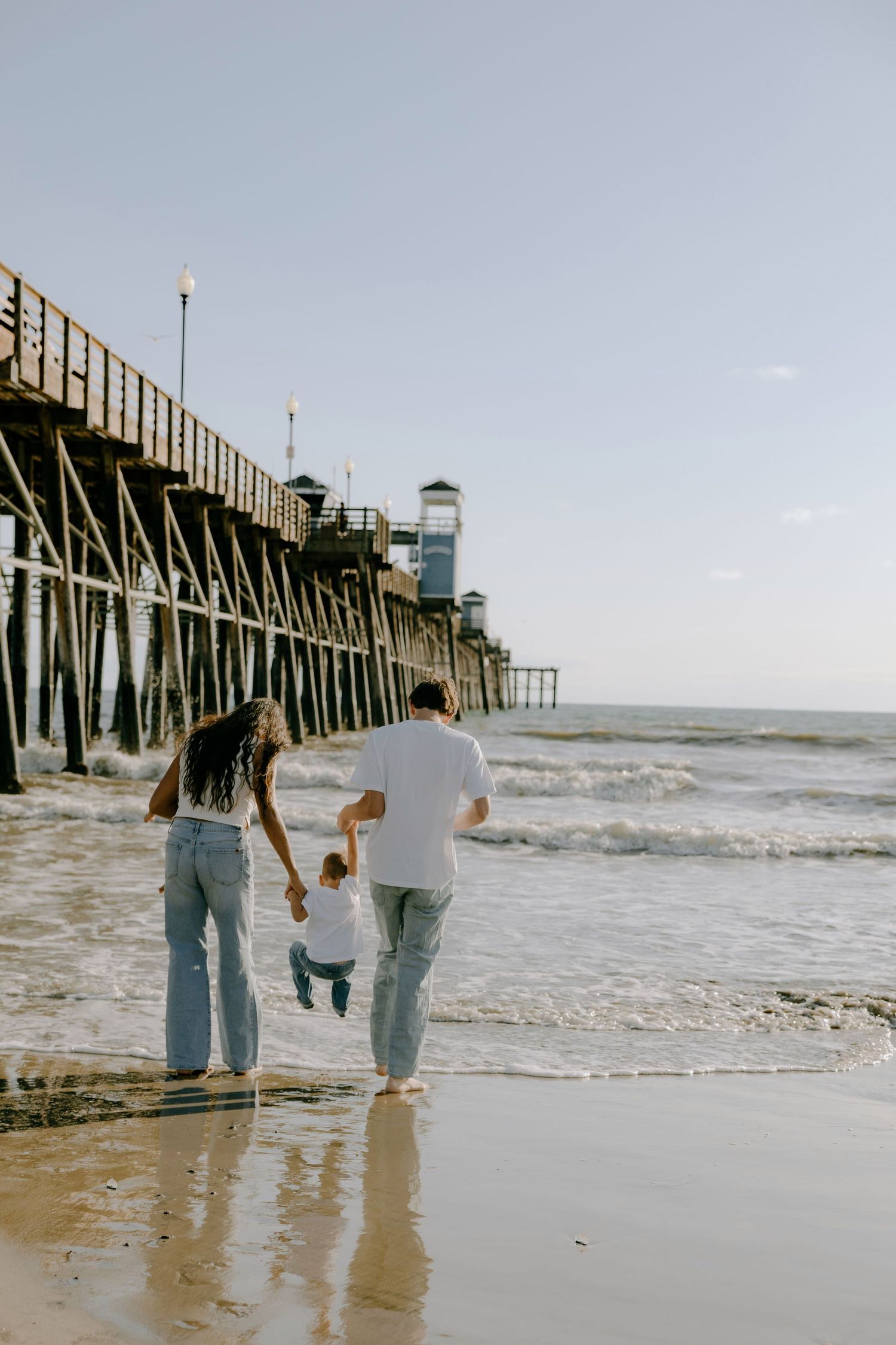 Family walking on beach near wooden pier, parents swinging toddler over ocean waves on sunny day.