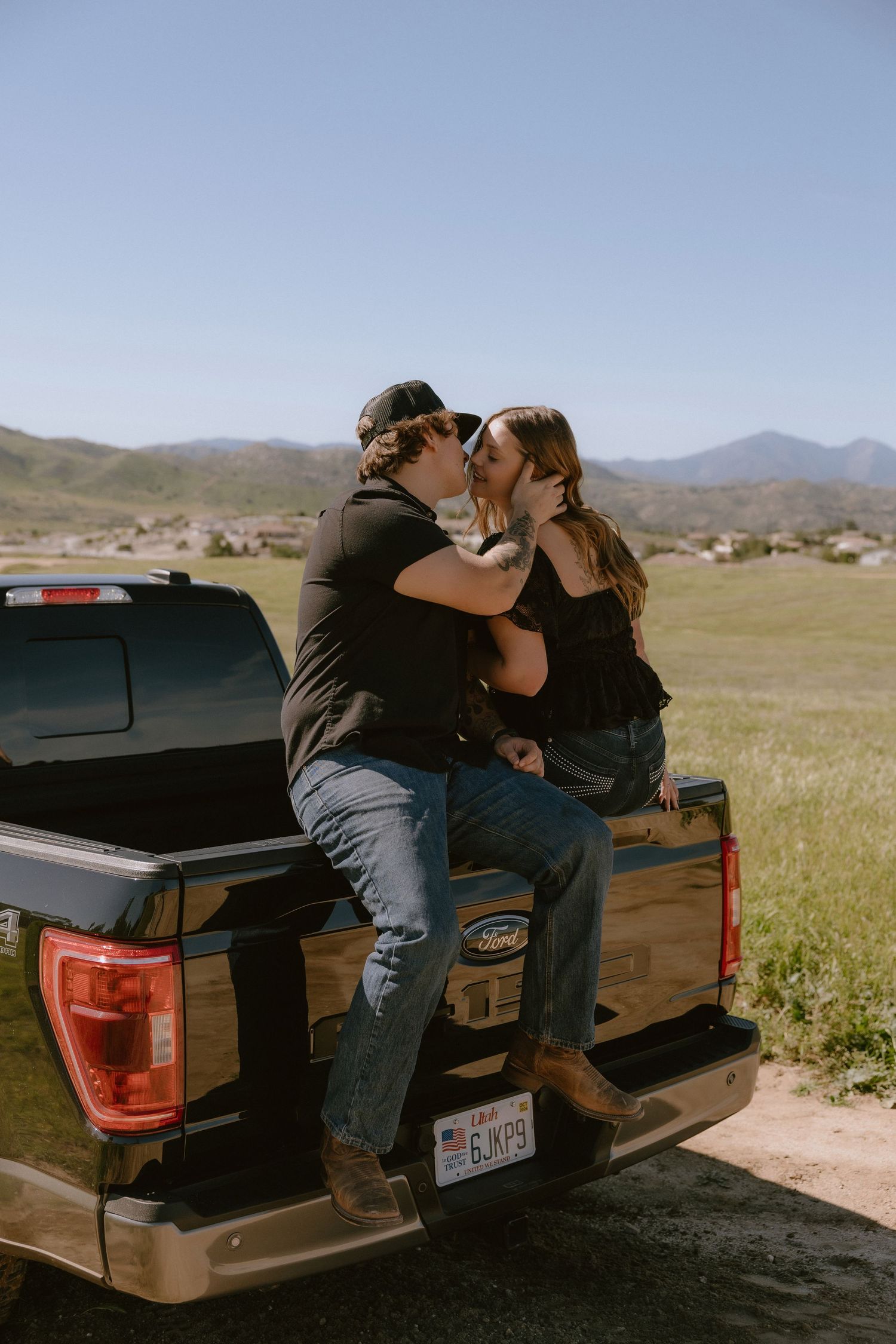 Two people sit in the back of a black pickup truck in a rural landscape with mountains in the background.