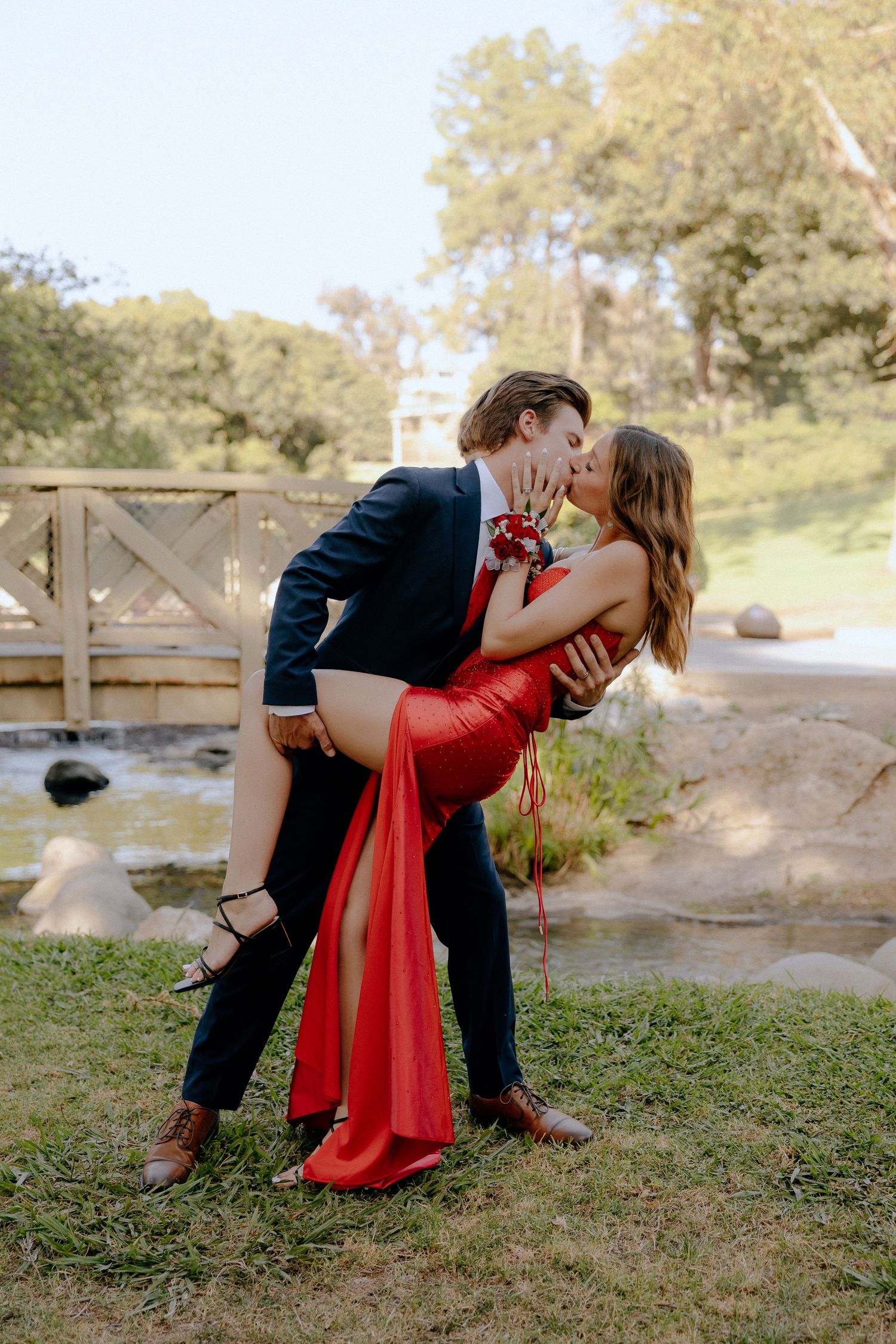 Couple sharing a romantic dip kiss outdoors, wearing red gown and navy suit near a wooden bridge.