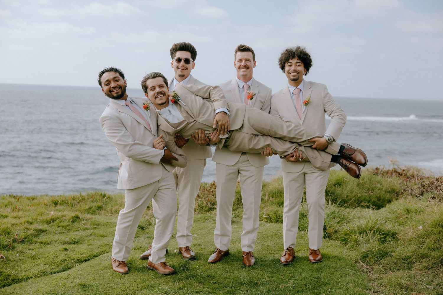Five groomsmen in tan suits celebrate outdoors near the ocean, lifting the groom horizontally while laughing.