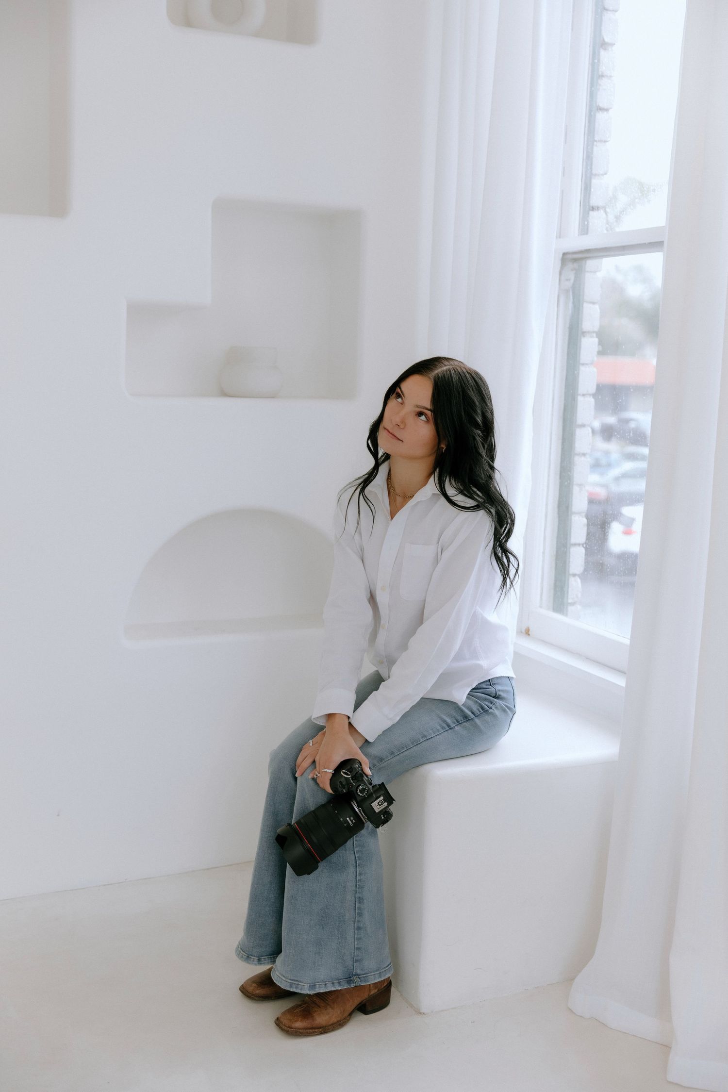 A photographer in a white sweater and jeans sits on a white window ledge holding a camera in a minimalist setting.