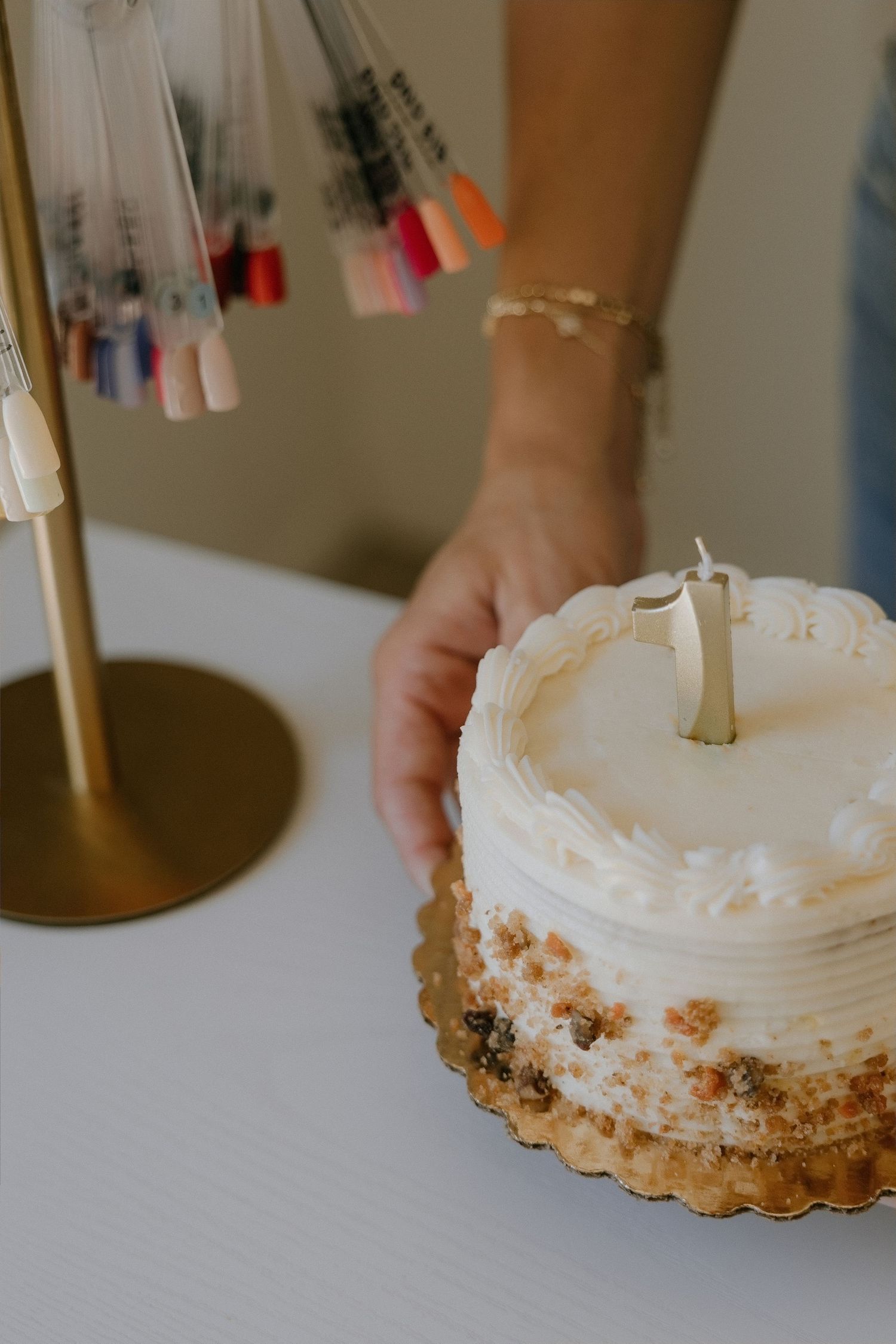 A hand with a gold bracelet places a white frosted cake with a candle on a table near a nail polish display.