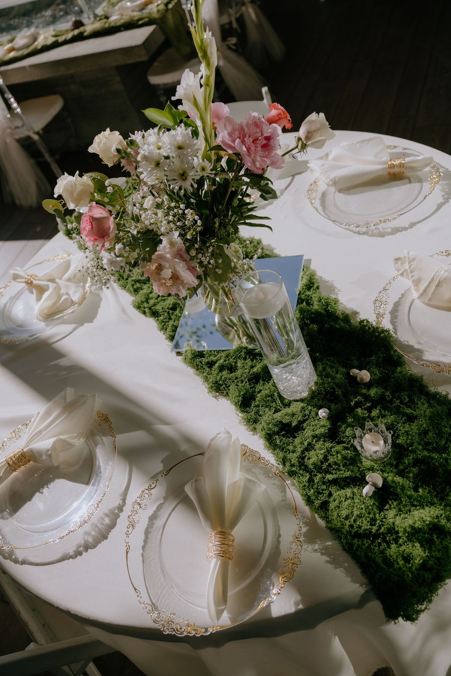 Elegant wedding table setting with moss runner, pink floral centerpiece, gold-rimmed plates, and white napkins on white tablecloth.