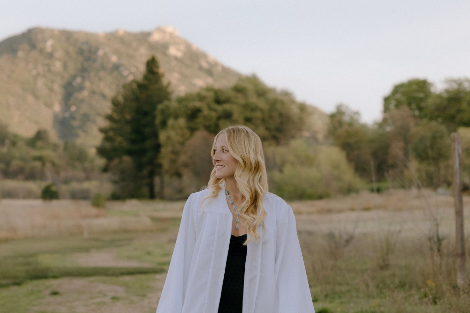 Blonde woman in white wrap standing in scenic mountain meadow with trees and green hills backdrop.