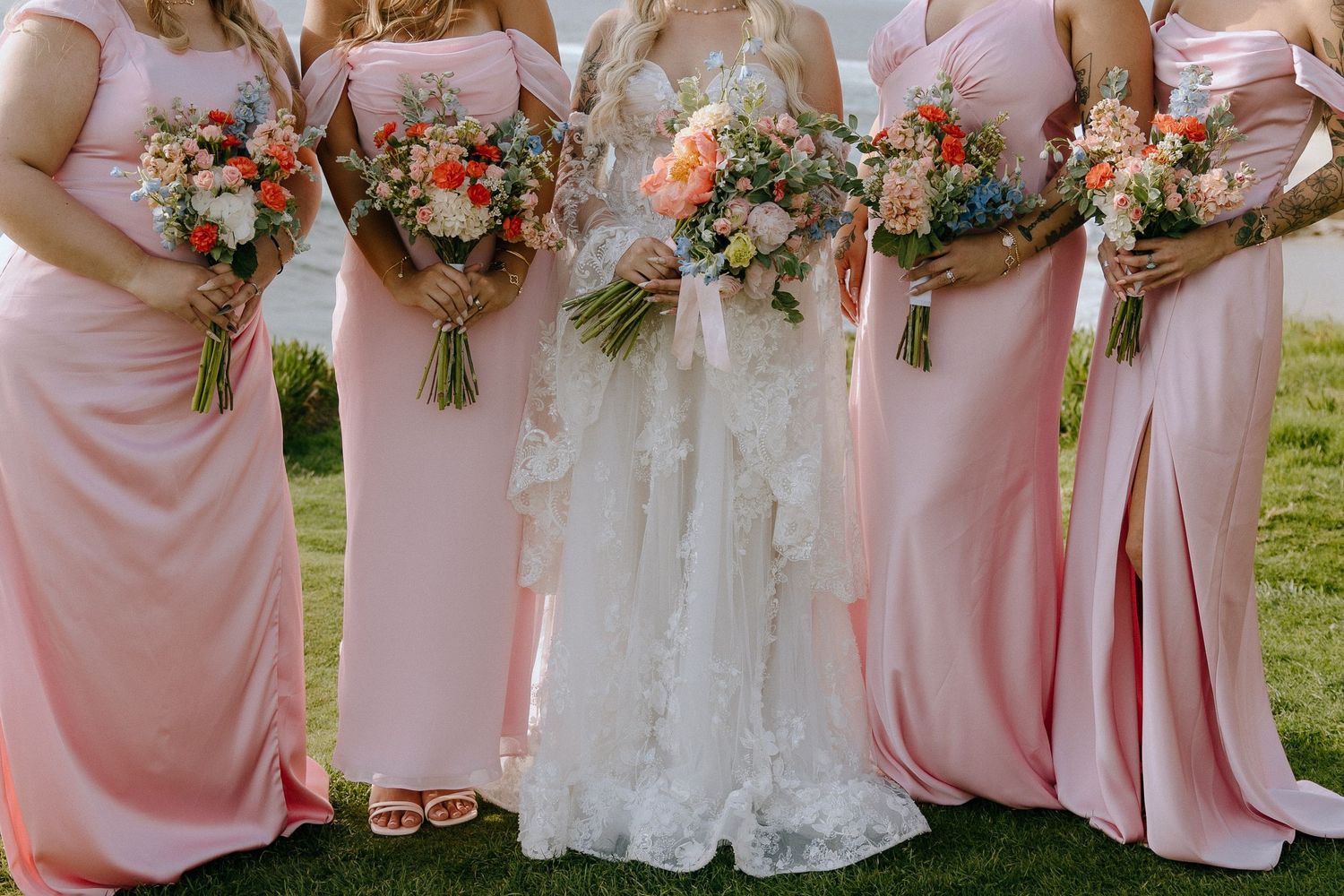 Bride in lace gown with bridesmaids in pink dresses holding colorful floral bouquets outdoors.