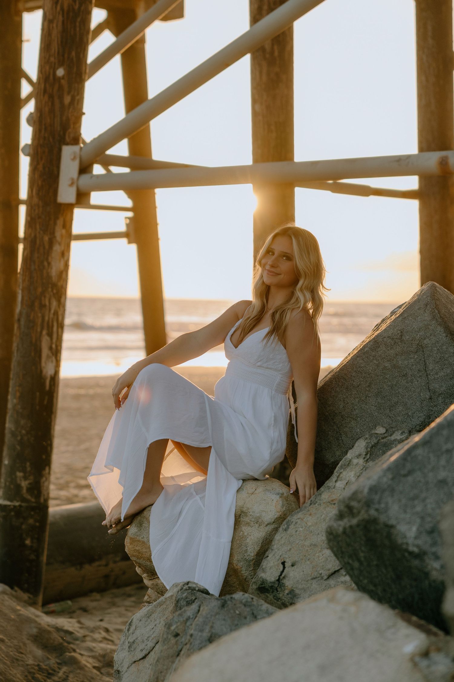 Blonde woman in white maxi dress sitting on rocks under pier at golden sunset by the ocean.