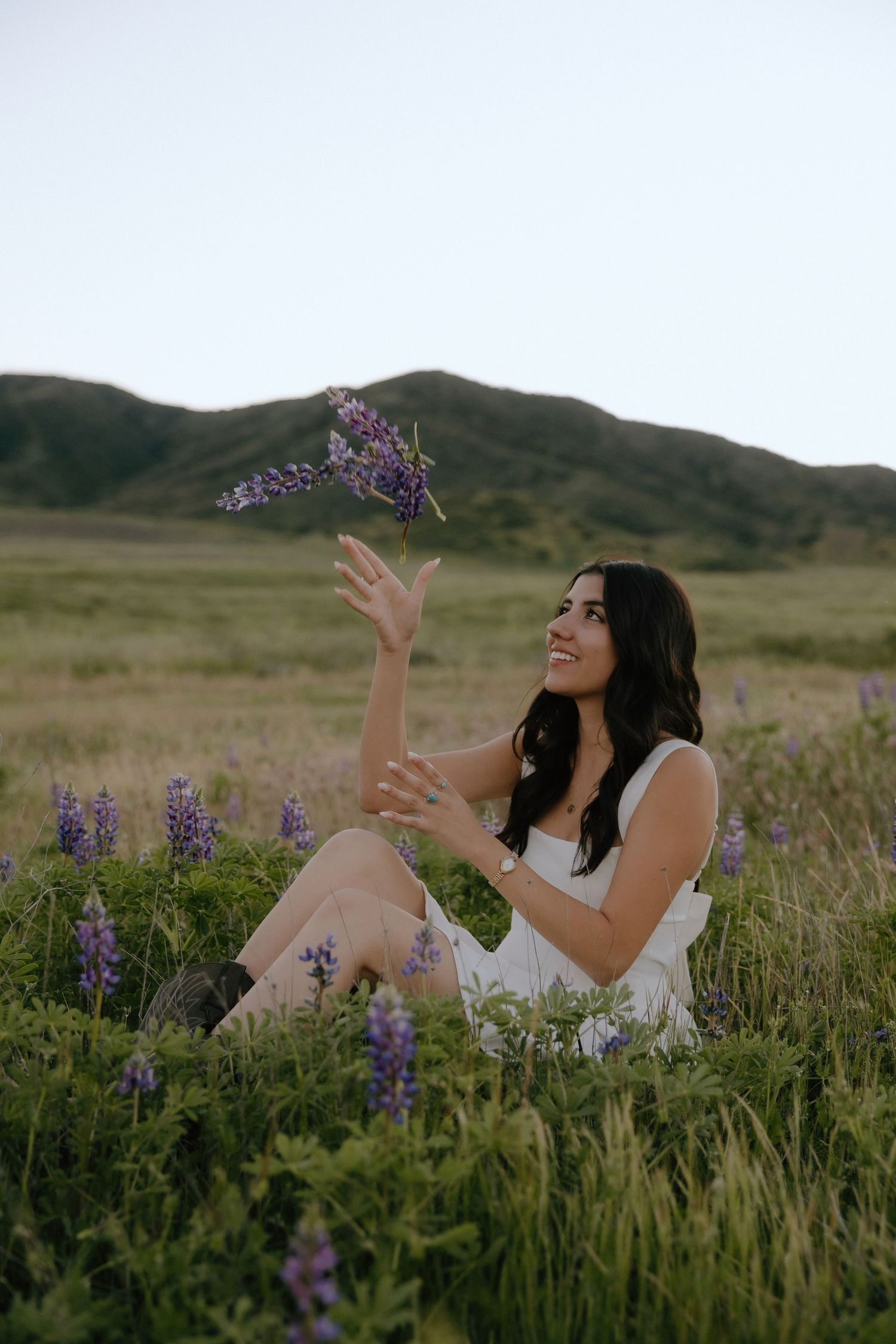 A smiling woman in a white dress sits in a lupine field, tossing purple wildflowers in the air with green hills behind.