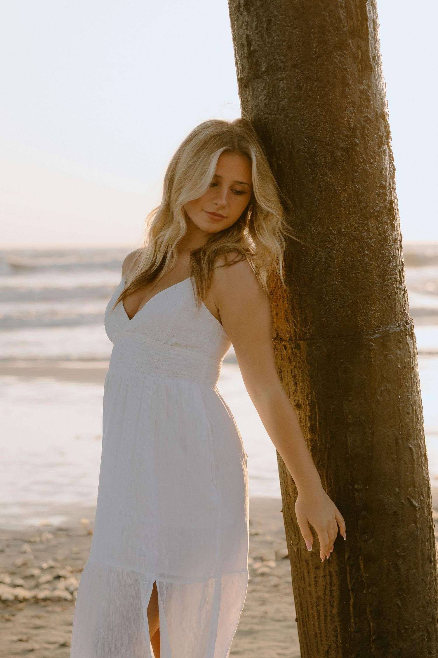 Blonde woman in white sundress leaning against a palm tree on a sandy beach at golden hour.