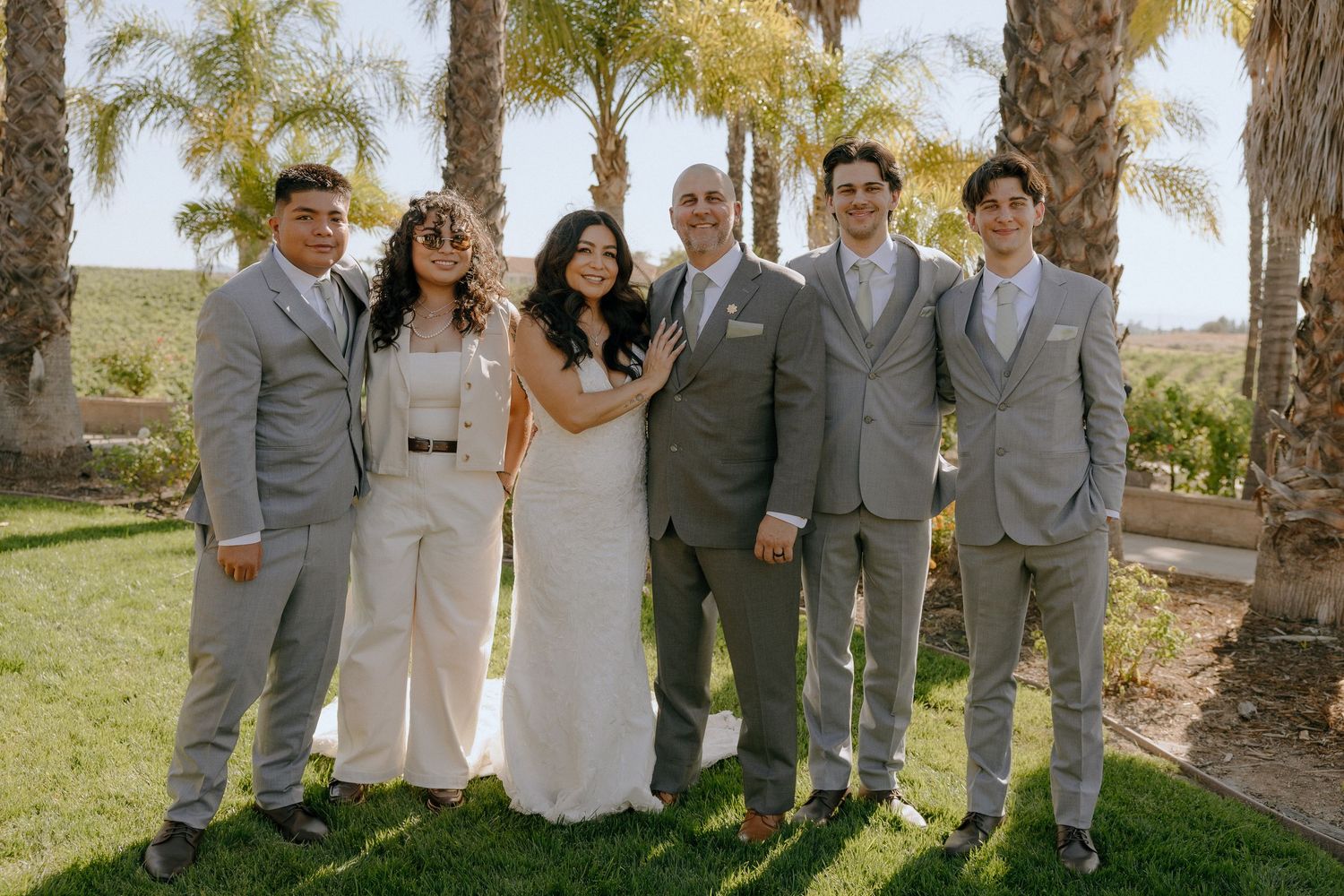 Wedding party poses outdoors on green lawn with palm trees, bride in white gown flanked by groom and groomsmen in gray suits.
