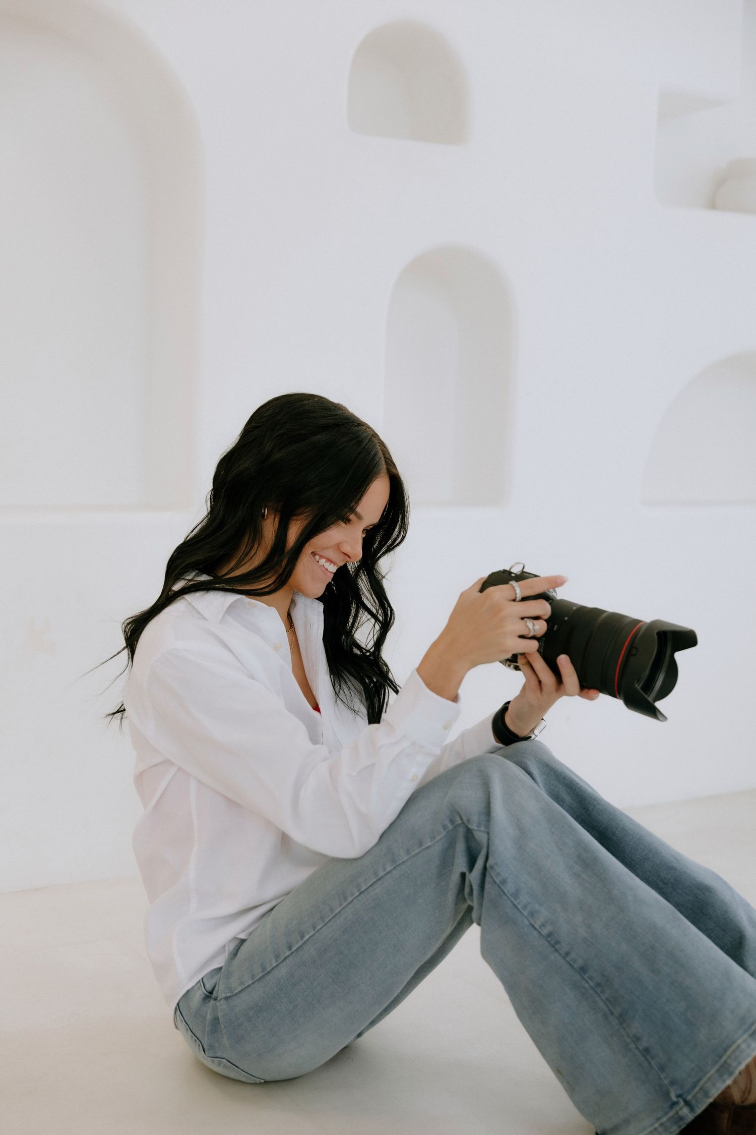 Photographer in white shirt and jeans reviews images on professional camera while sitting against white wall with arches.