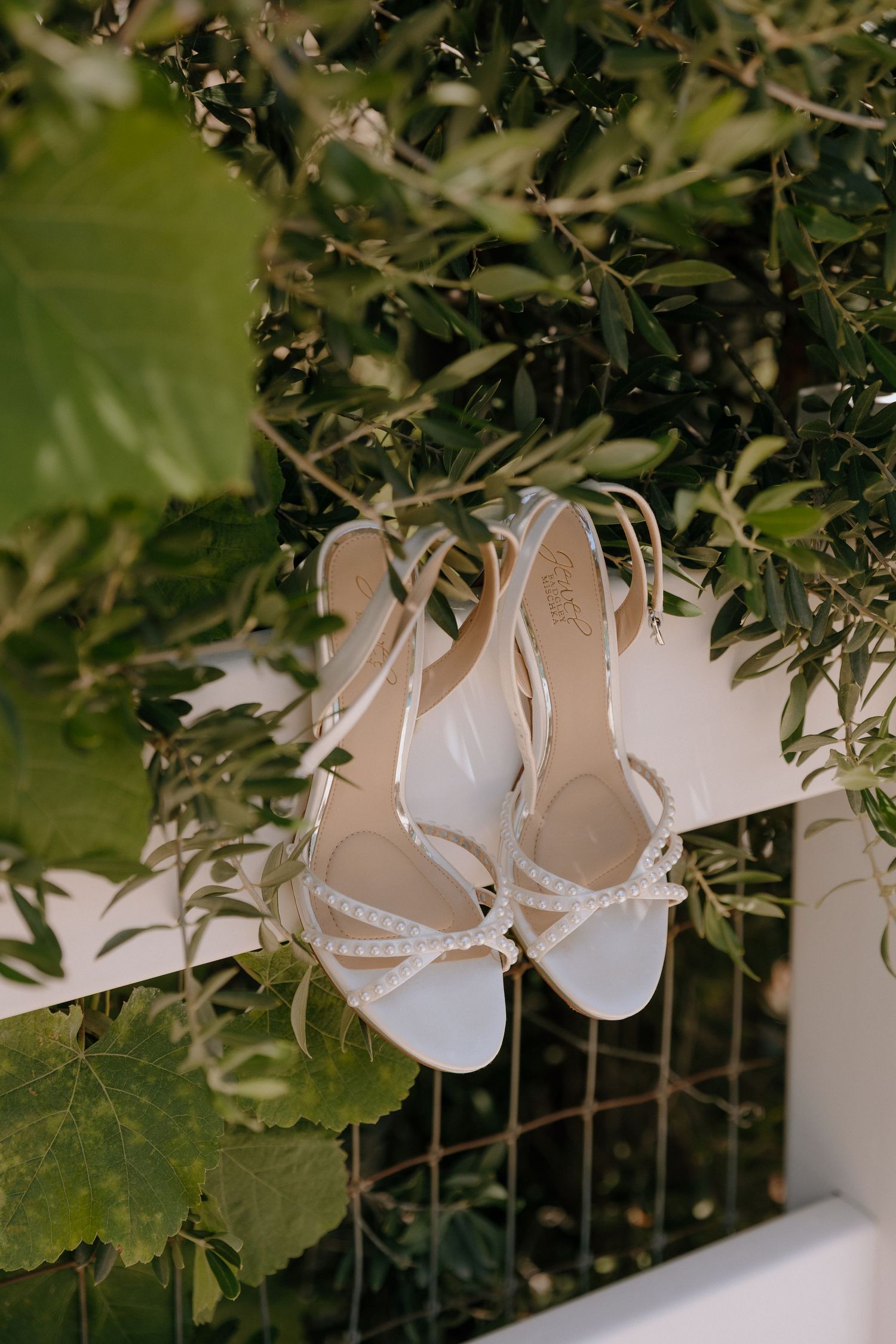 White bridal heels with pearl embellishments displayed on a white fence surrounded by lush green foliage.
