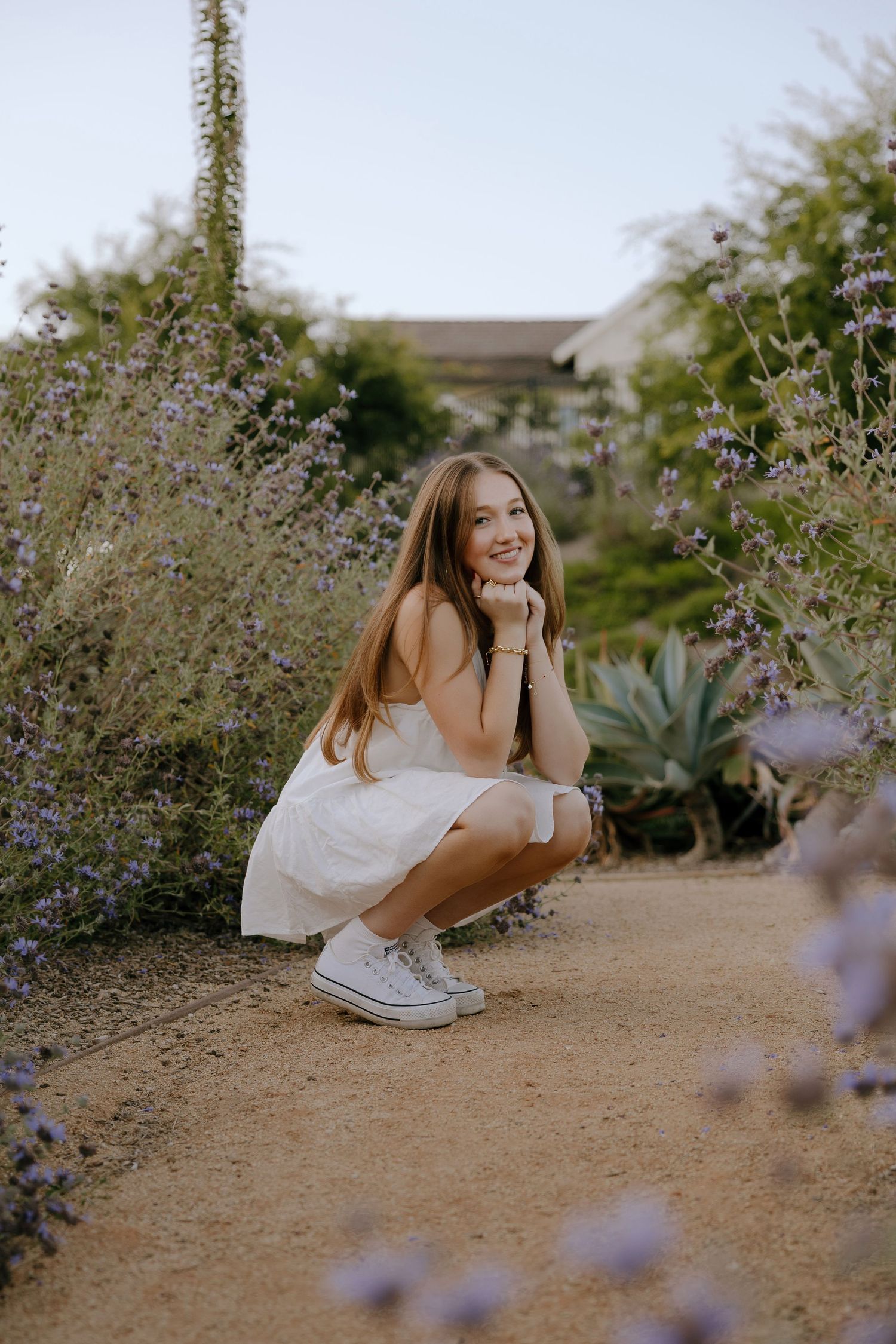 Young woman in white dress and Converse sneakers crouching on a garden path surrounded by purple wildflowers.
