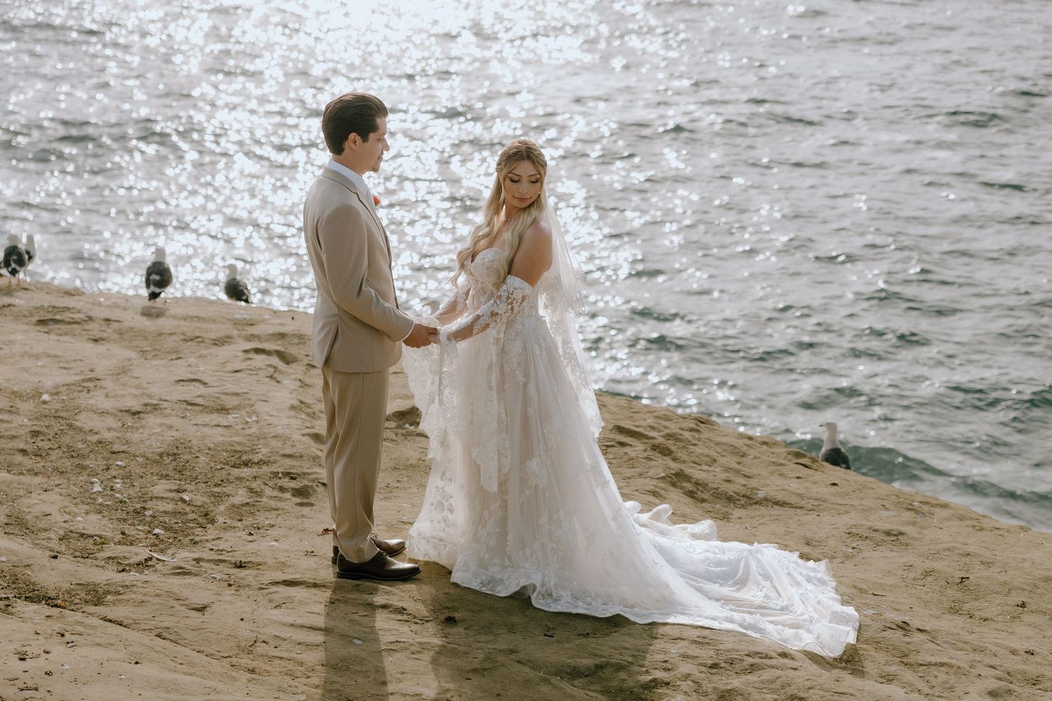 Bride and groom holding hands on sandy beach near sparkling water in elegant wedding attire.