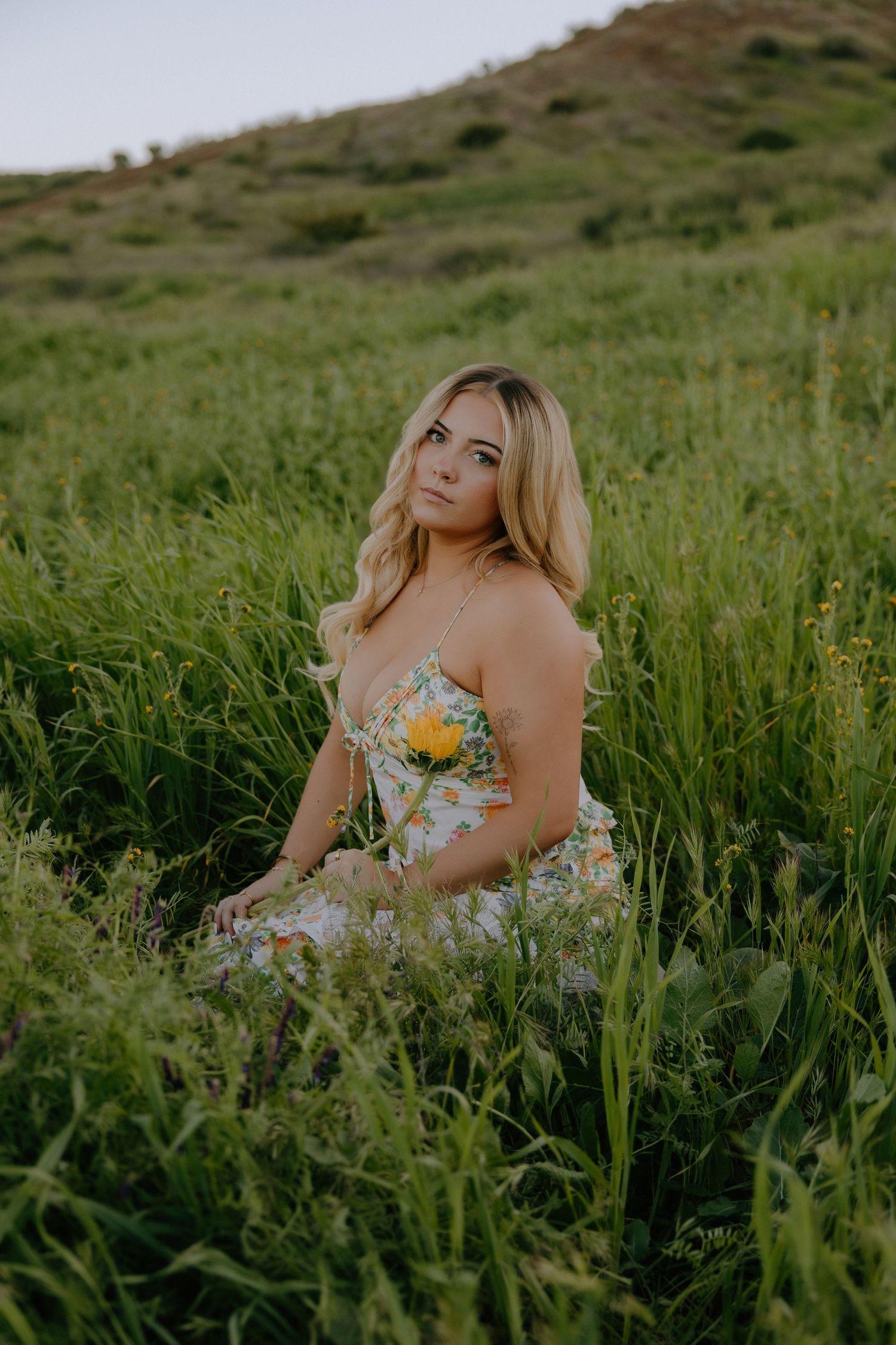 A woman in a floral dress poses gracefully in a field of tall grass on a hillside during golden hour.