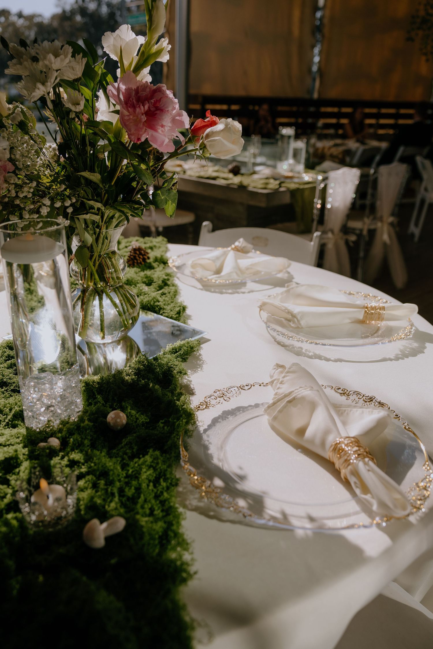 Elegant wedding table setting with gold-rimmed plates, white napkins, moss runner, and floral centerpiece with pink and white blooms.