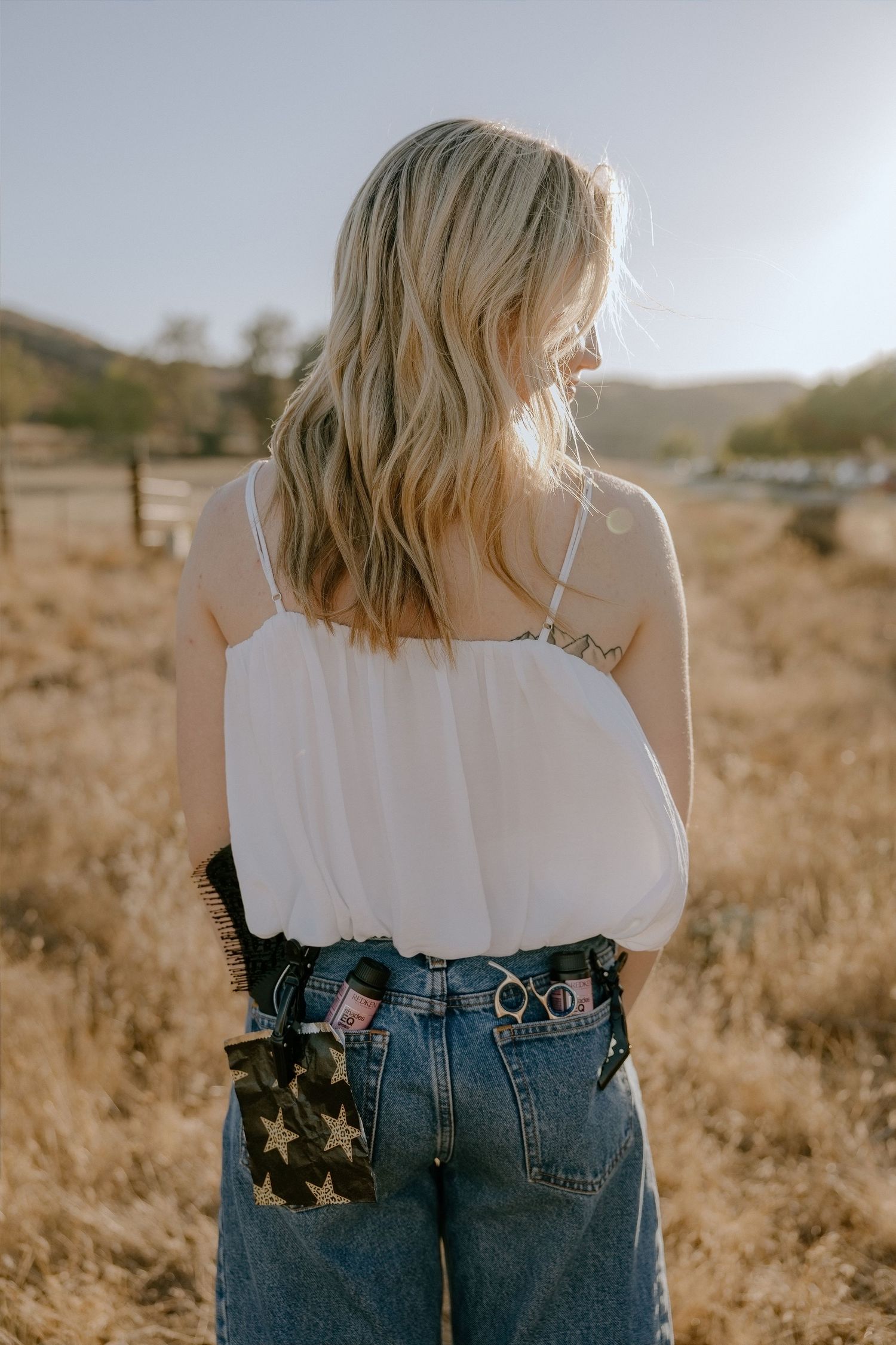 Blonde woman in white cami top and star-print denim jeans standing in a golden field during sunset.