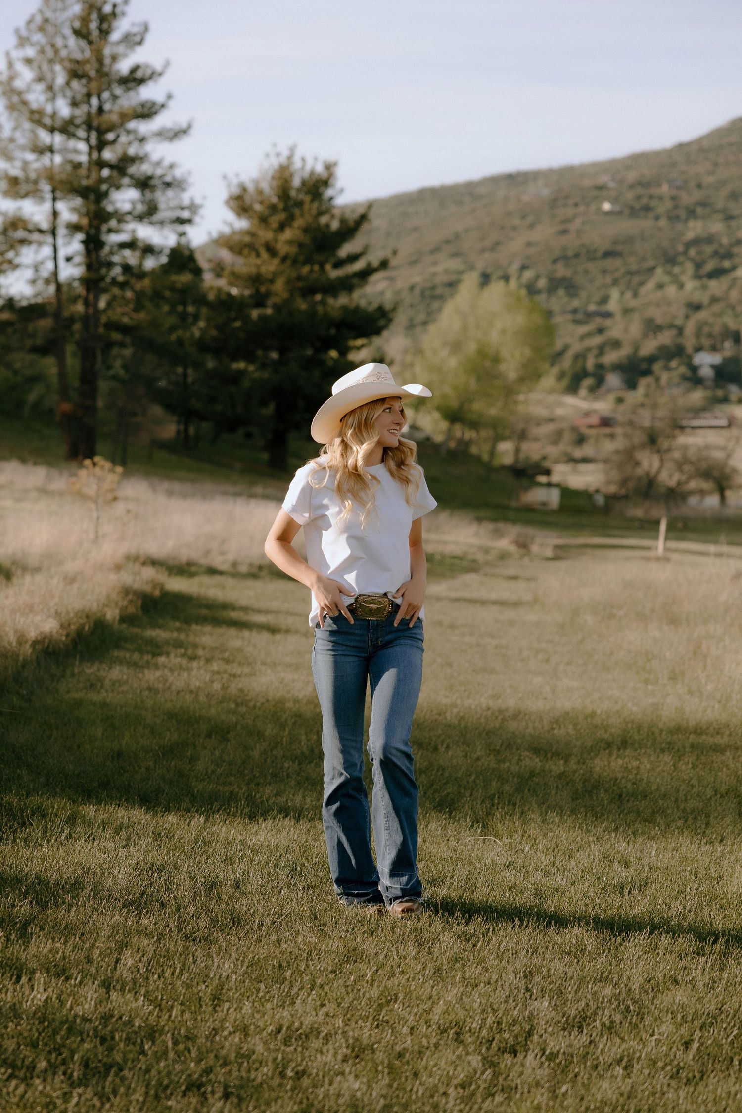 Woman in white cowboy hat, white tee, and flare jeans standing in a sunny green field with hills behind her.