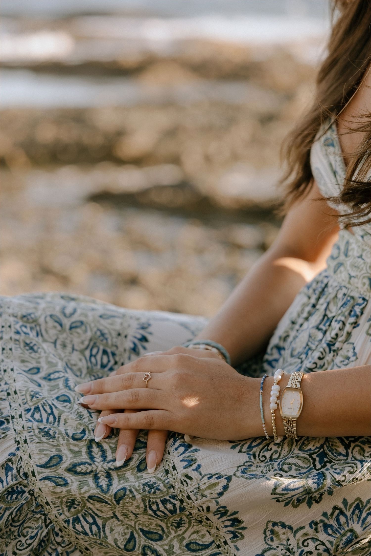Woman in boho floral dress wearing gold bracelets and ring, sitting on rocky beach in warm sunlight.
