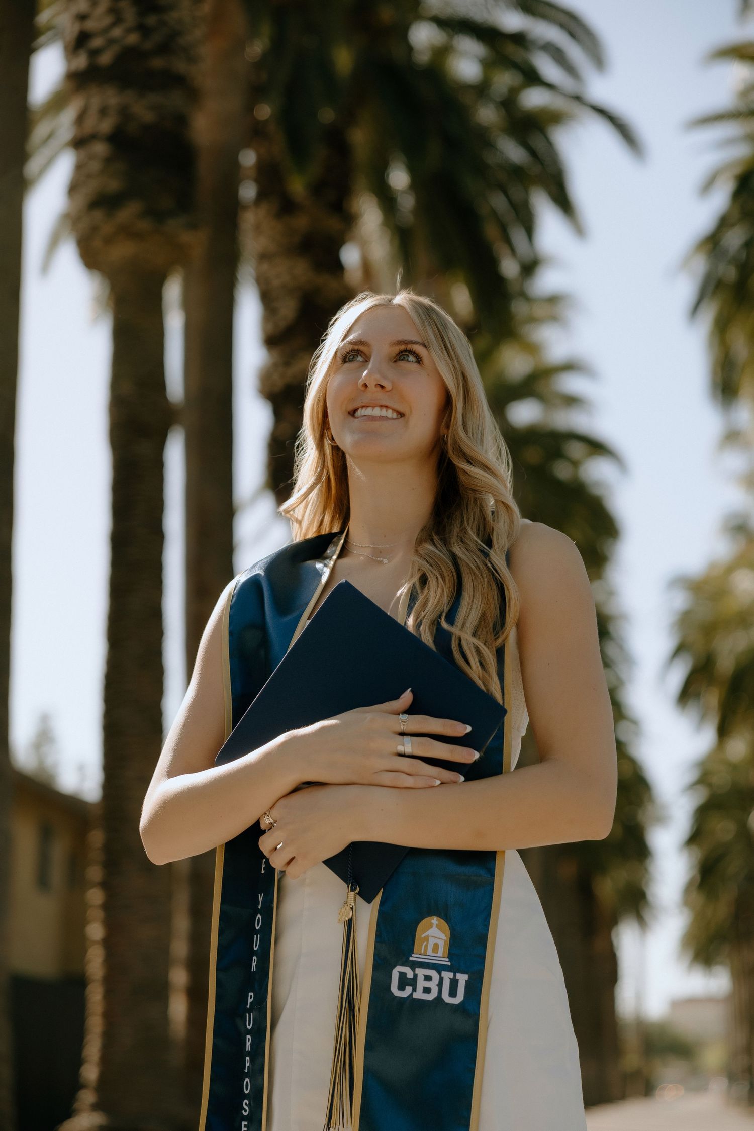 A smiling CBU graduate holds her diploma while wearing a blue sash outdoors among tall palm trees.