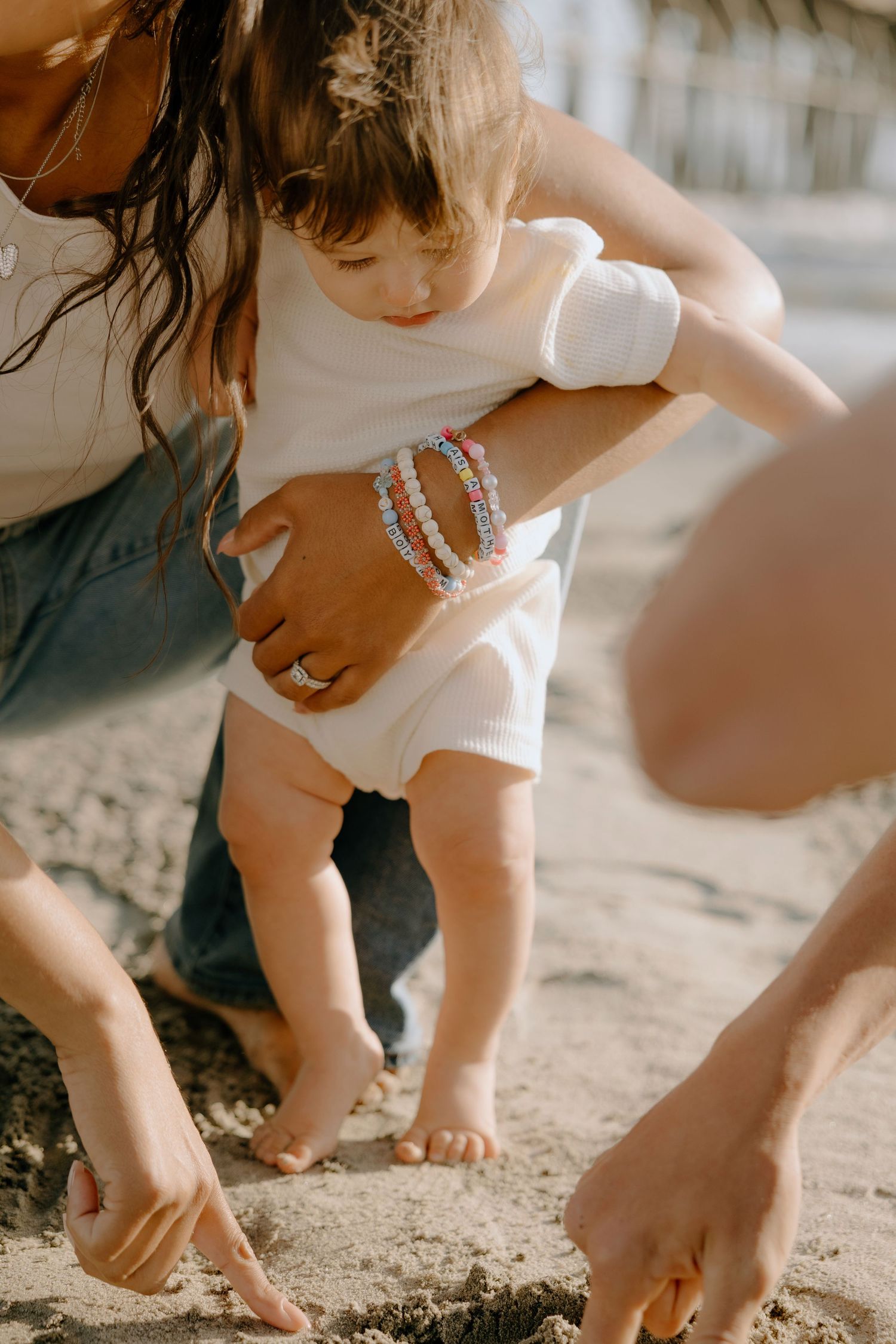 Mother helping toddler stand on sandy beach, wearing colorful beaded bracelets and a white onesie.