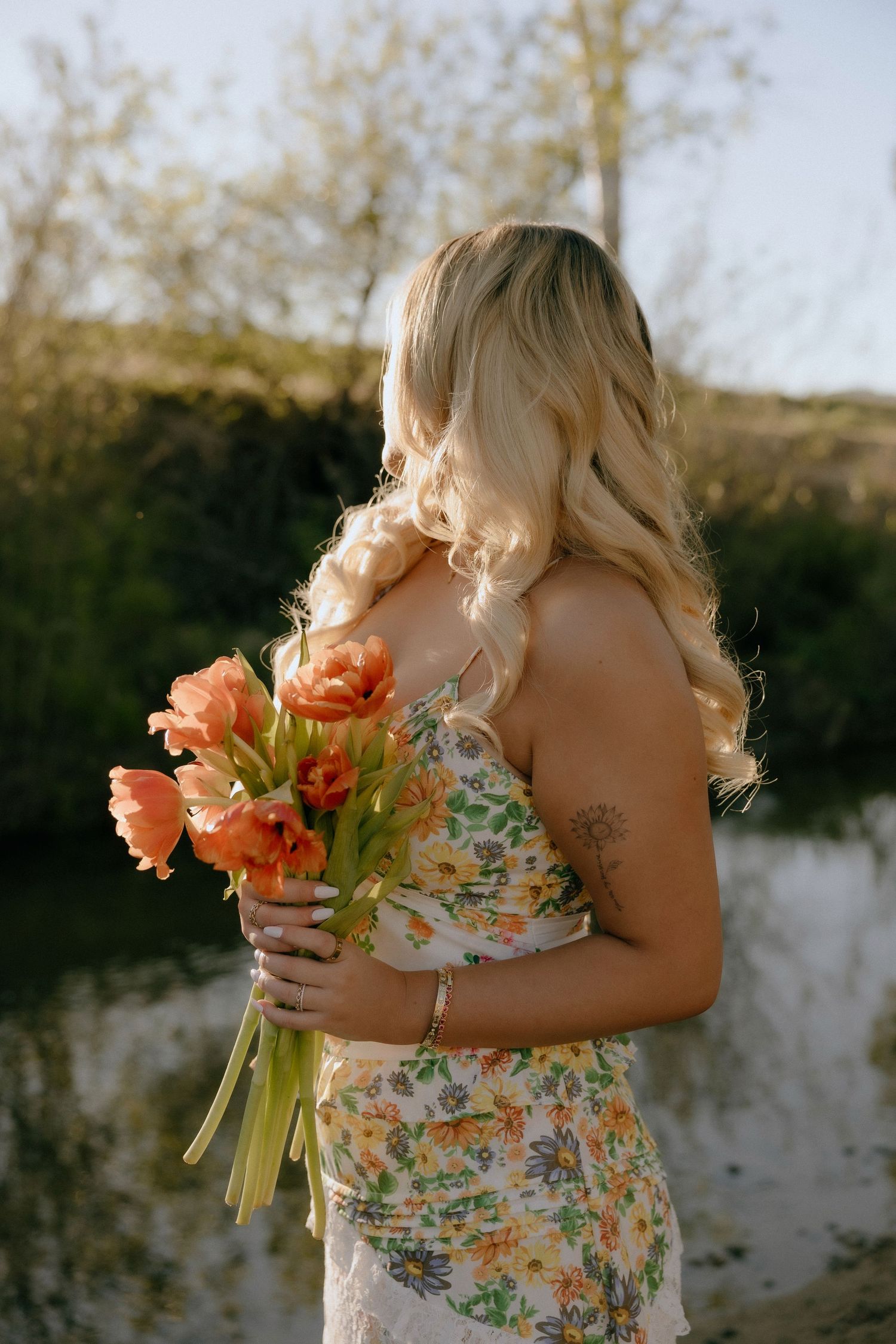A person in a floral dress holds vibrant orange tulips by a lake at sunset, with soft golden light illuminating their hair.