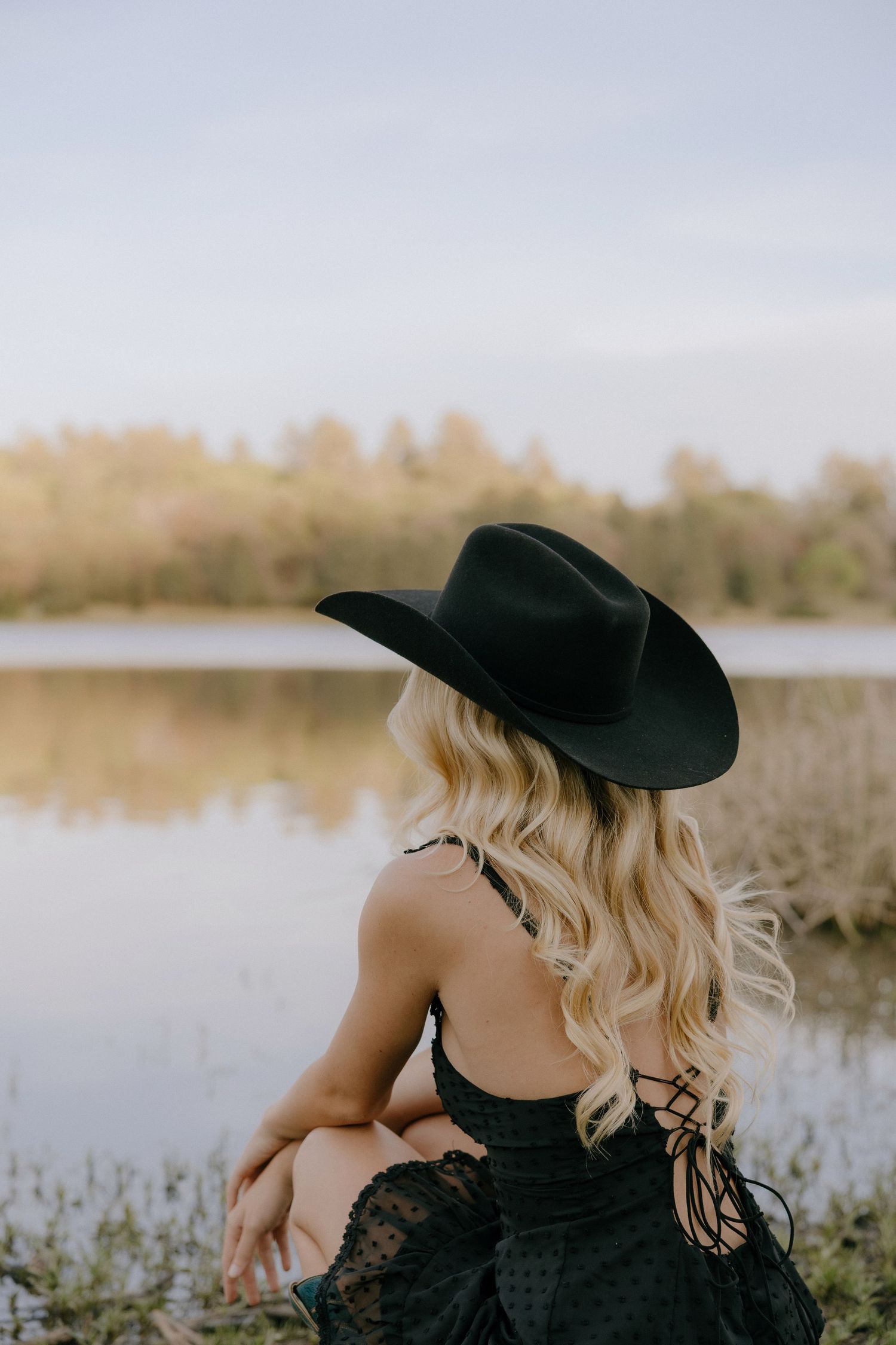 Blonde woman in black cowboy hat and lace dress sitting by a serene lake with autumn trees reflected in water.