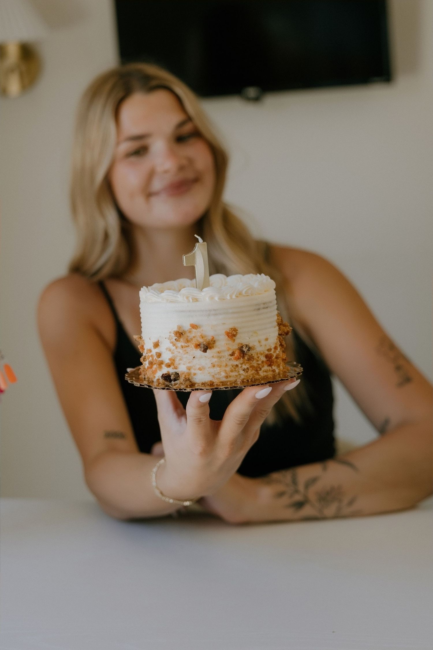 Smiling blonde woman with tattoos holding a white frosted birthday cake with a number one candle on top.