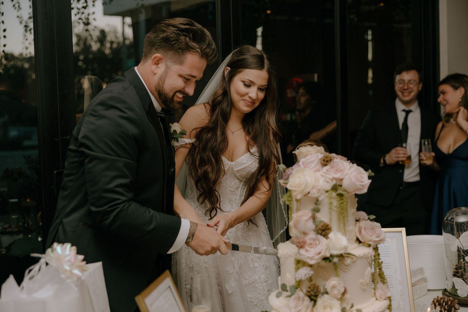 Bride and groom cutting wedding cake together at elegant reception, surrounded by guests celebrating in background.