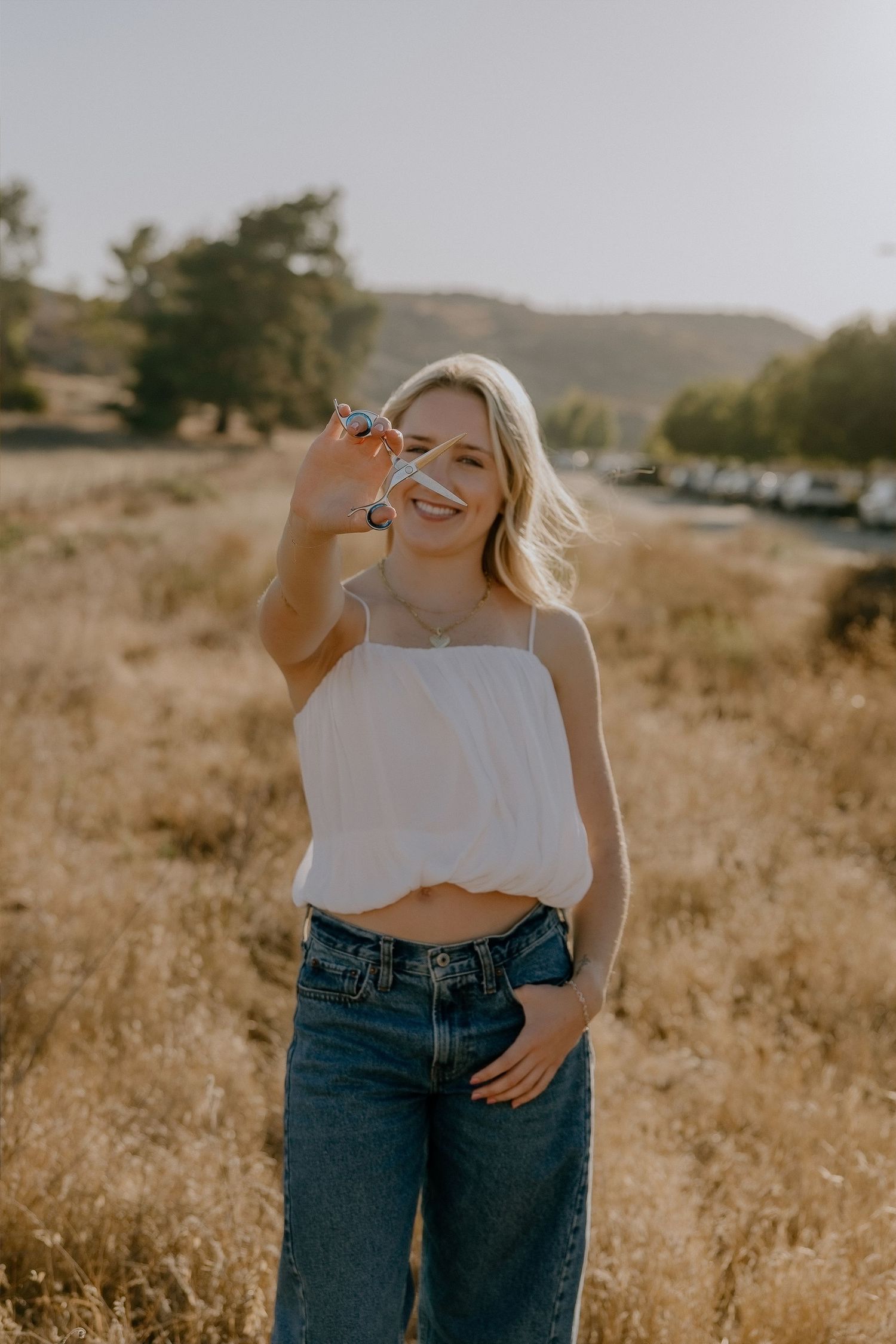 Smiling blonde woman in white crop top and jeans holding crystal in sunny golden field.