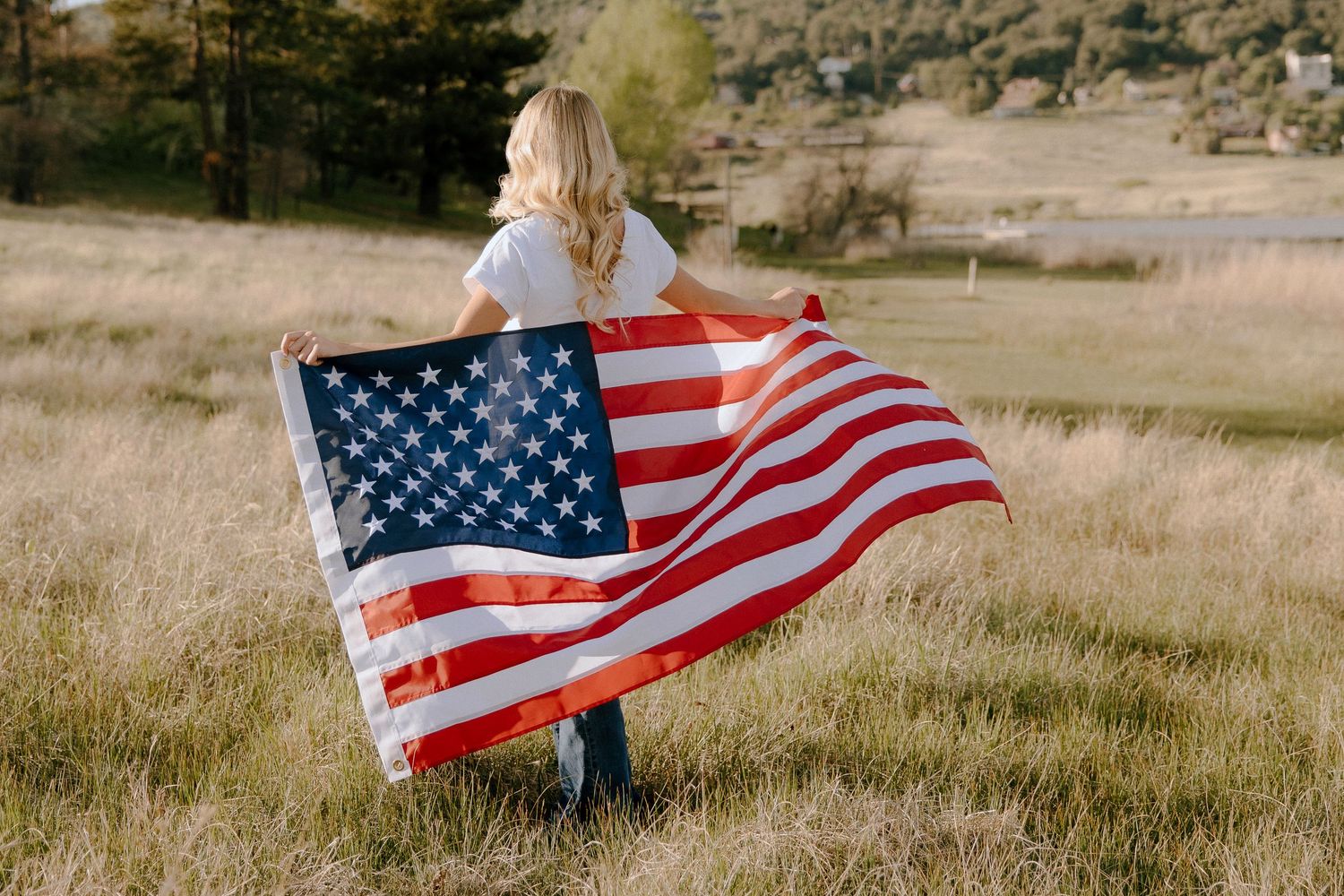 Blonde woman holding a large American flag in a golden grass field outdoors.