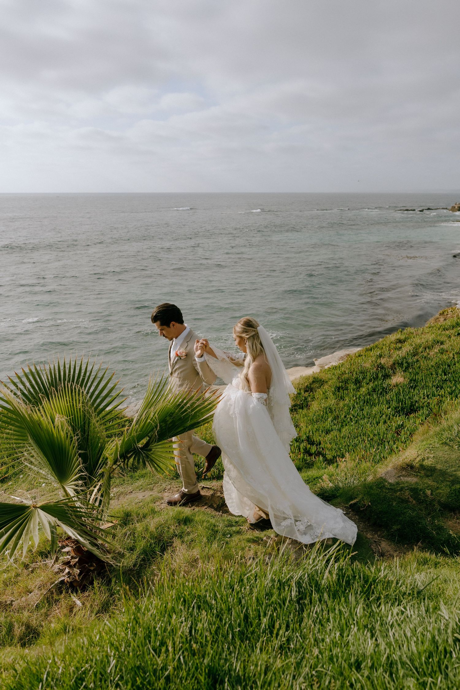 Bride and groom hold hands on a lush coastal cliff overlooking the ocean on their wedding day.