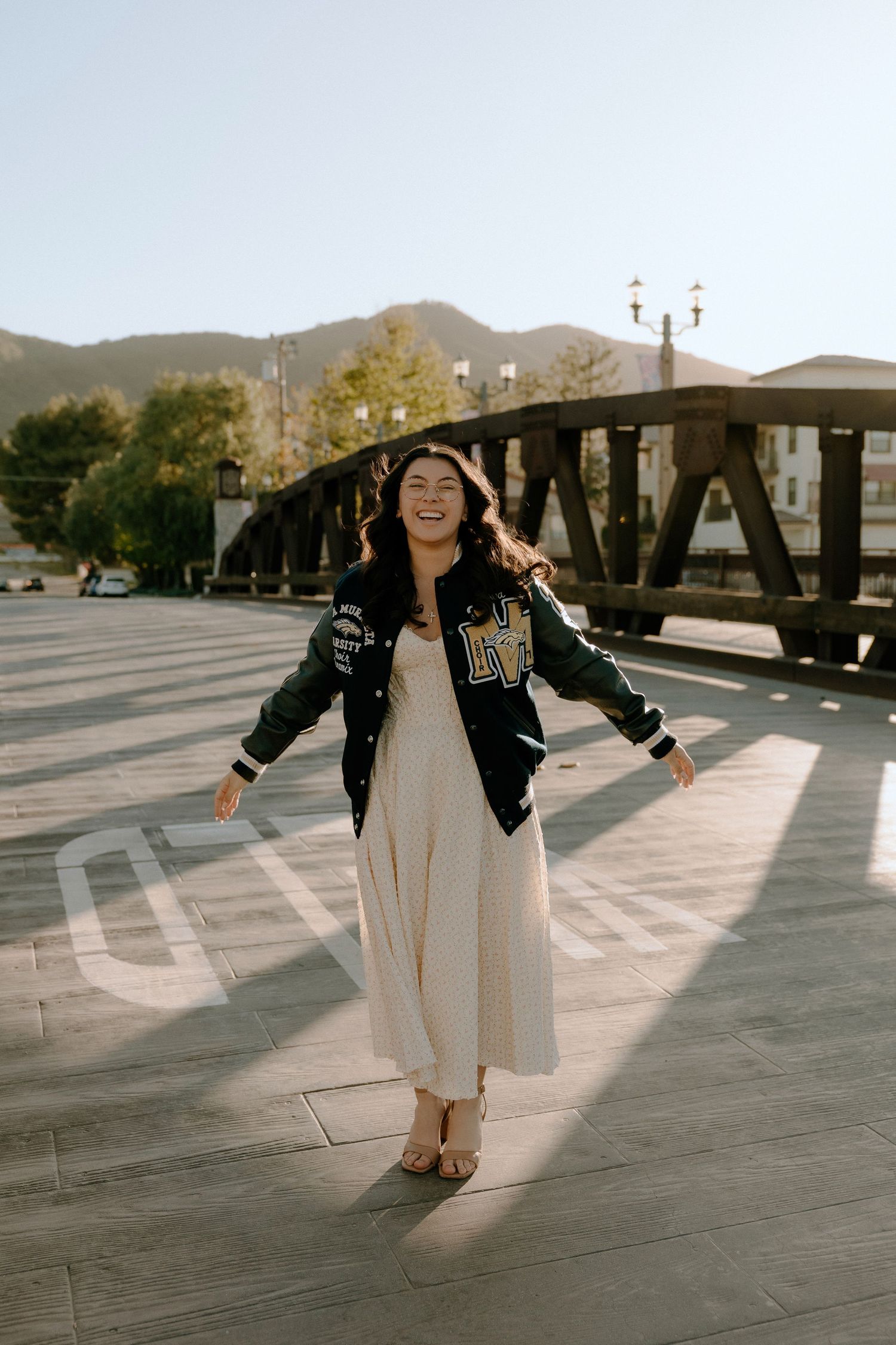 Smiling young woman in varsity jacket and cream dress twirling outdoors near a bridge on a sunny day.