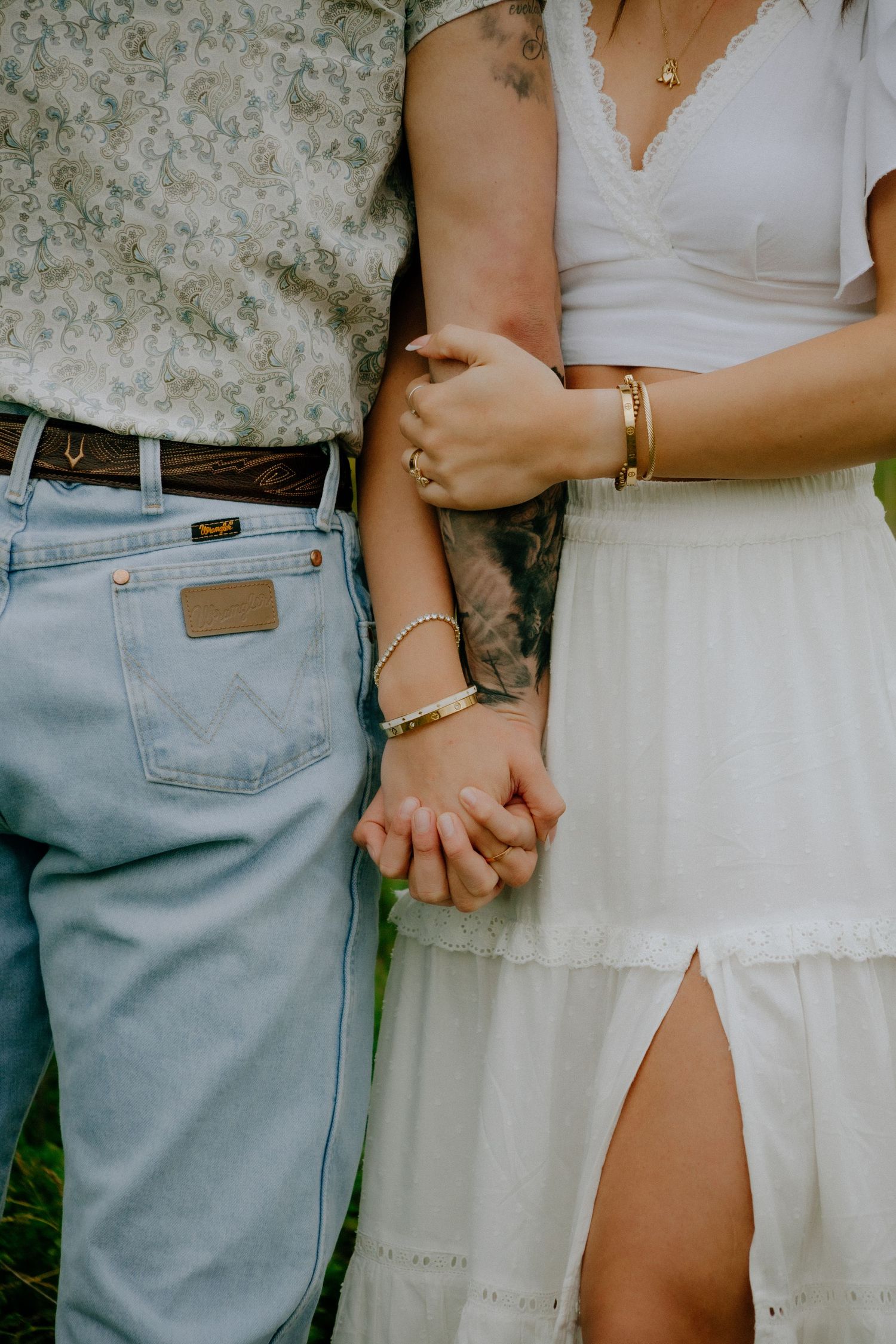 Close-up of couple holding hands, wearing contrasting light denim jeans and white summer dress with bracelets.