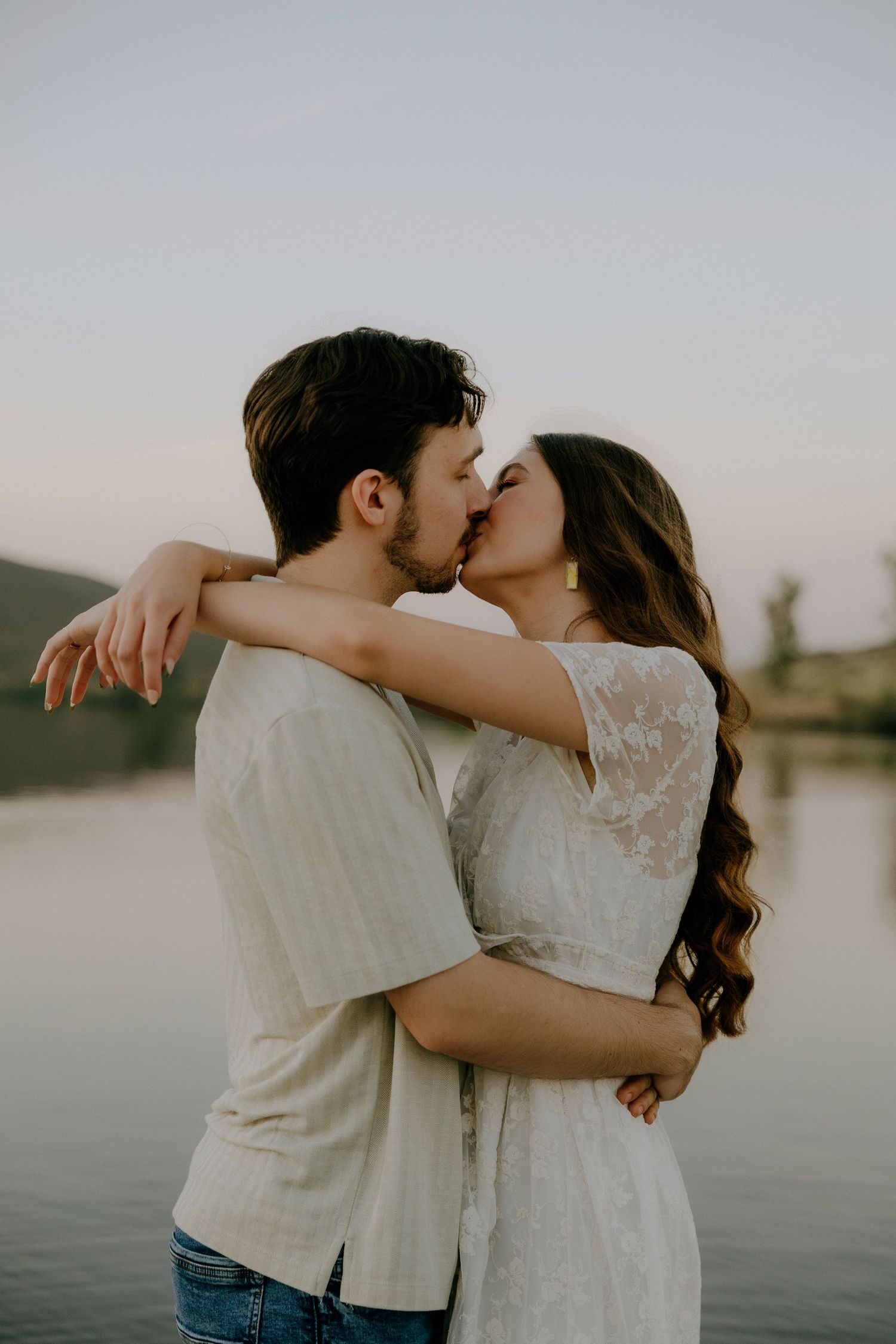 A couple shares a romantic kiss by a serene lake at sunset, embracing tenderly in soft neutral tones.