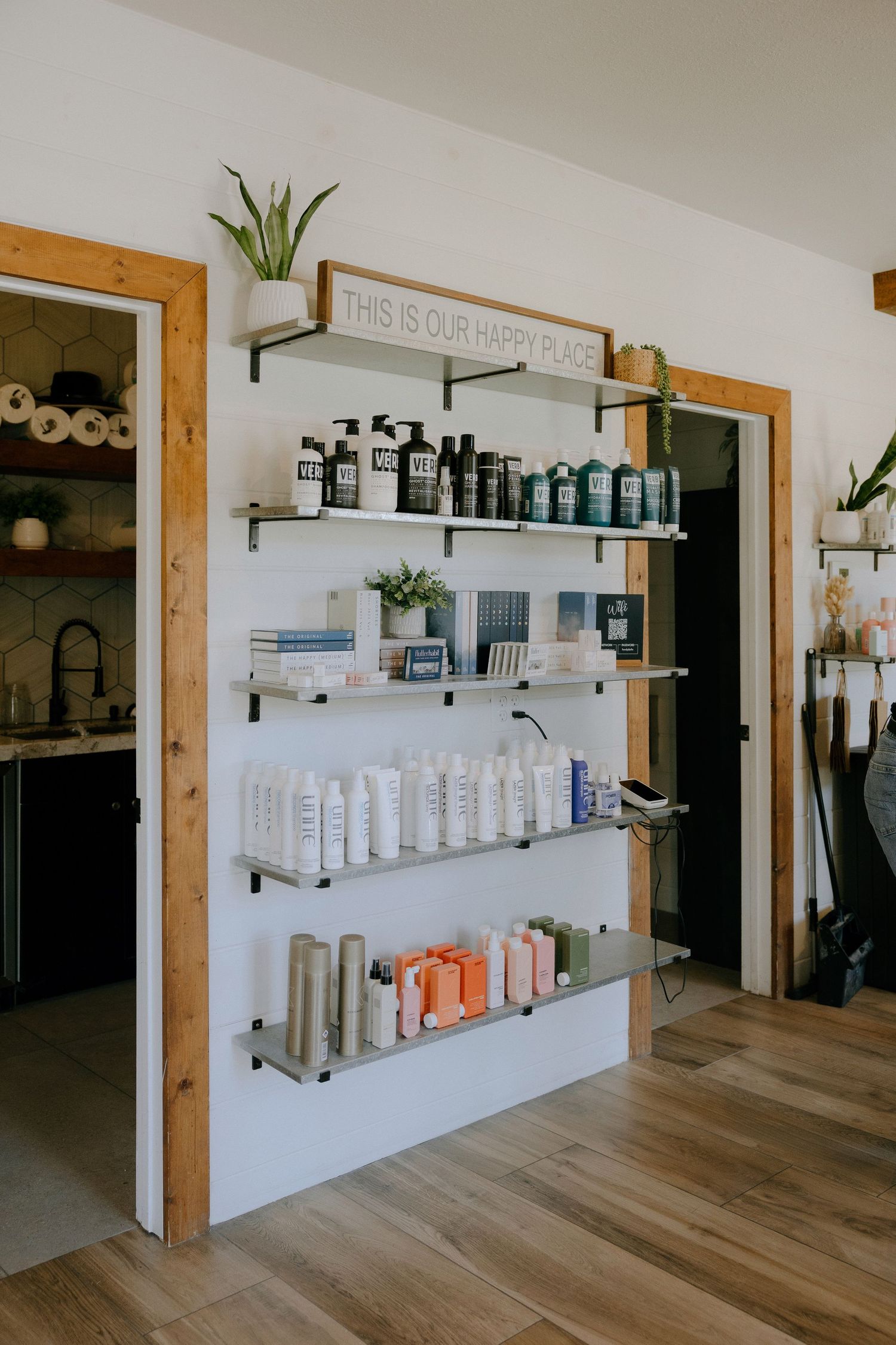Wall-mounted shelves displaying hair care products in a bright salon with hardwood floors and a This Is Our Happy Place sign.