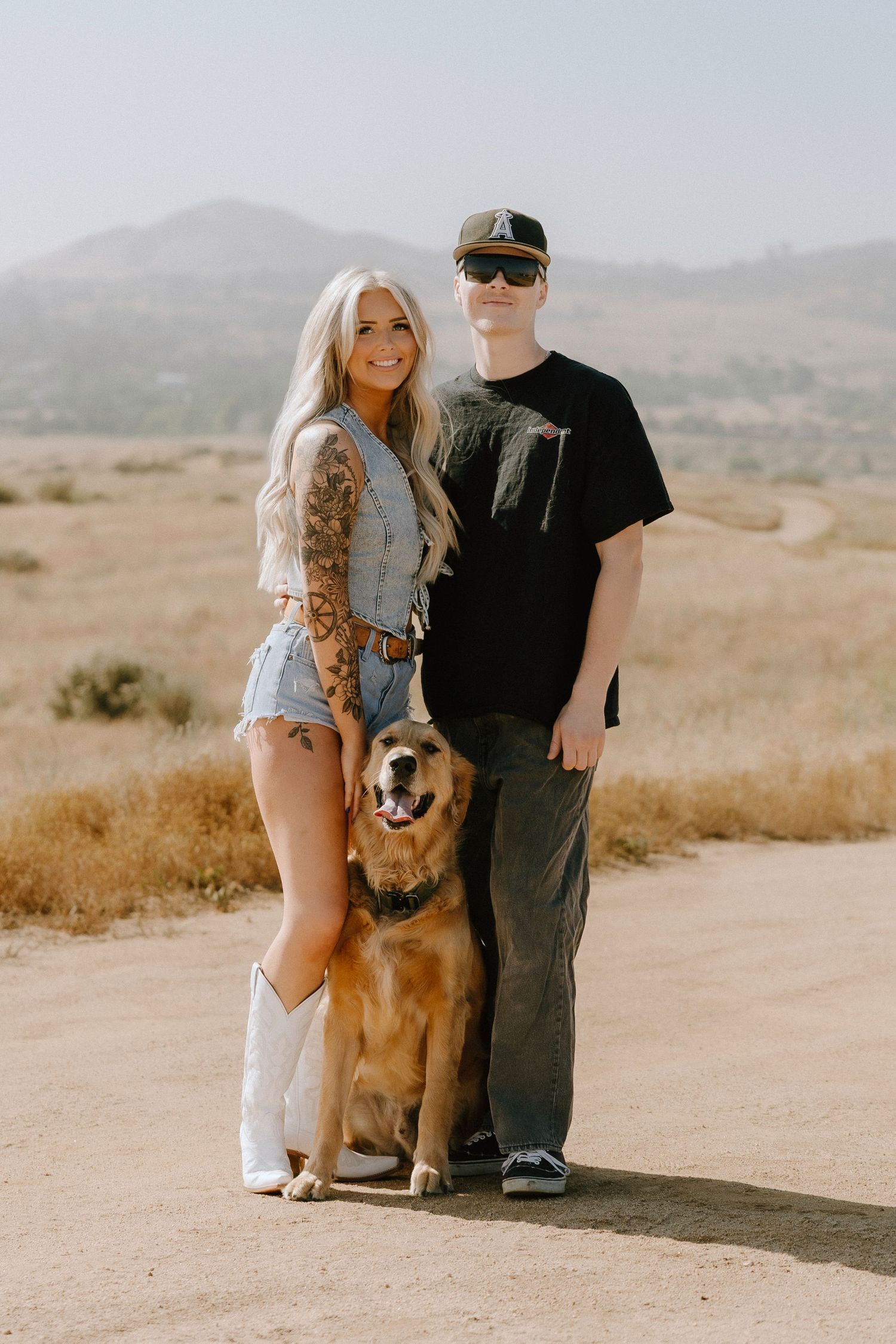 A couple and their golden retriever dog pose together on a dirt path with rolling hills in the background.