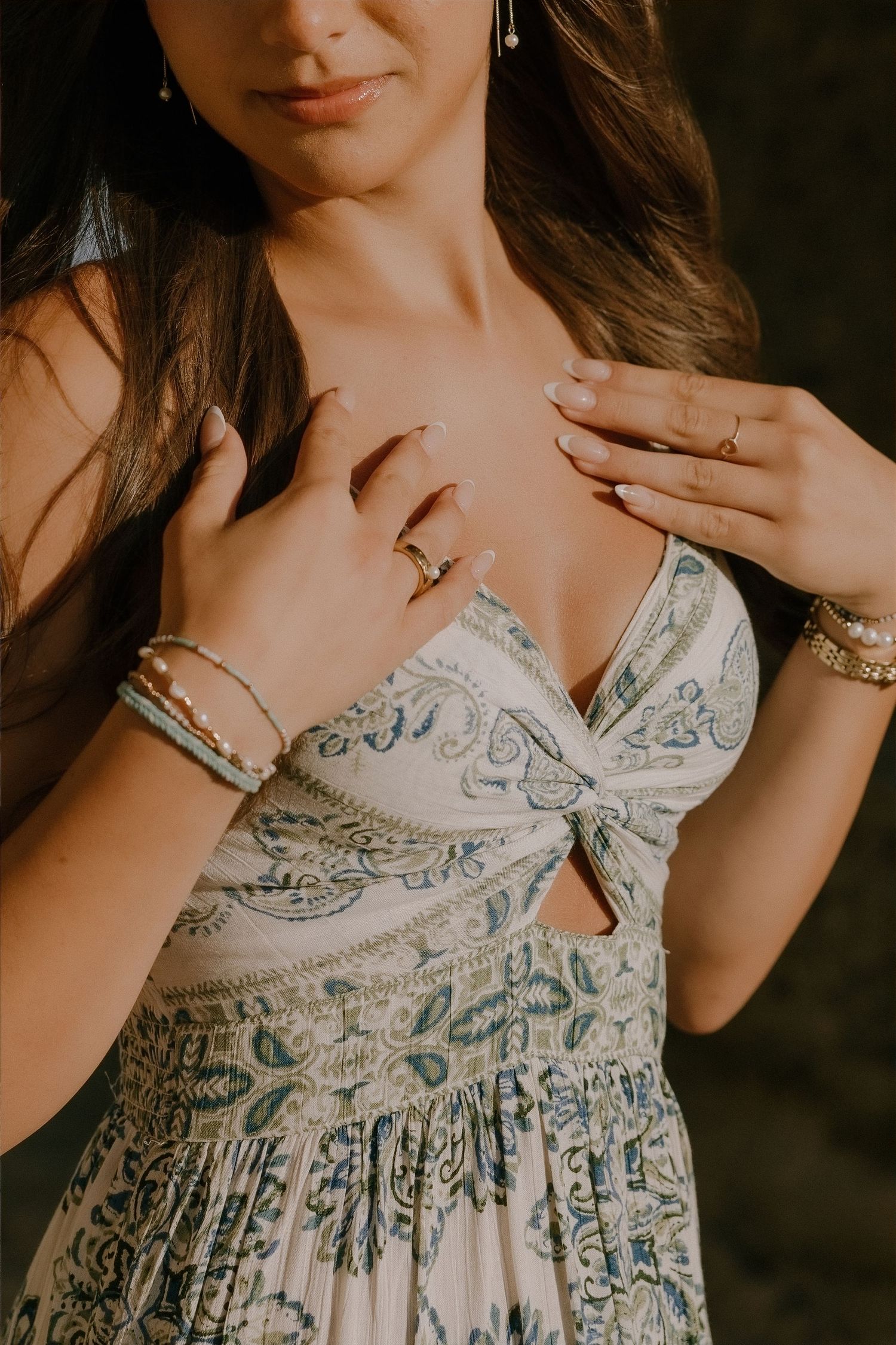 Woman in blue paisley dress wearing gold rings, bracelets, and earrings, posing elegantly with hands on chest.