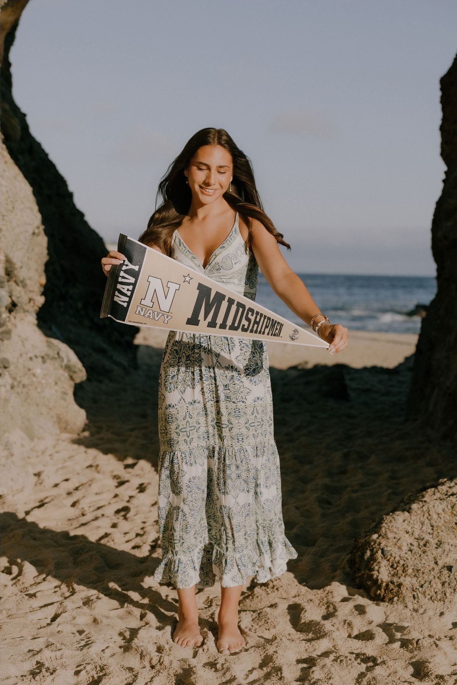 Young woman in floral dress holds Navy Midshipmen pennant flag on sandy beach near rocky cliffs.
