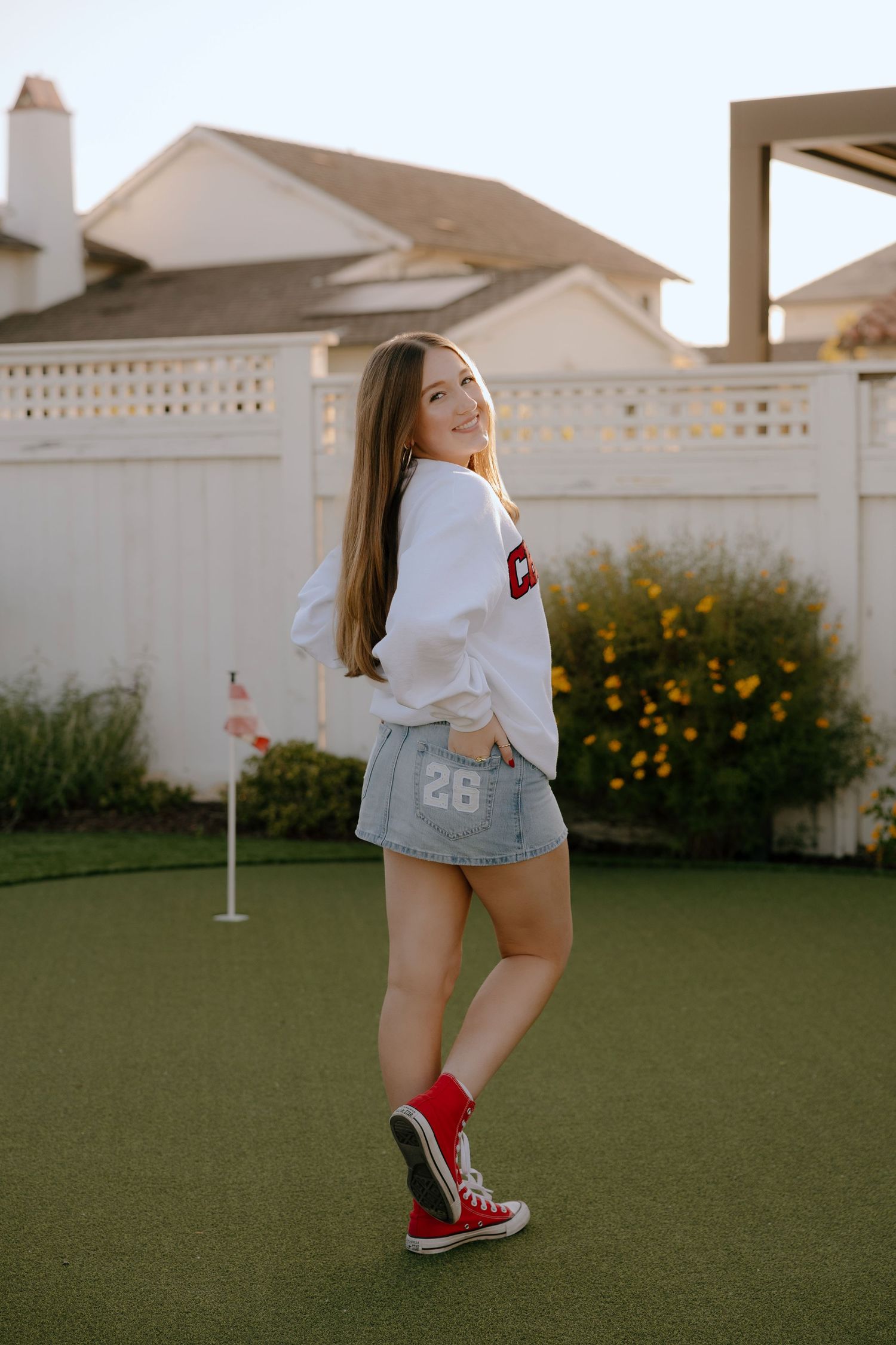Smiling teenage girl in white varsity sweatshirt, denim skirt, and red Converse sneakers posing on a backyard putting green.