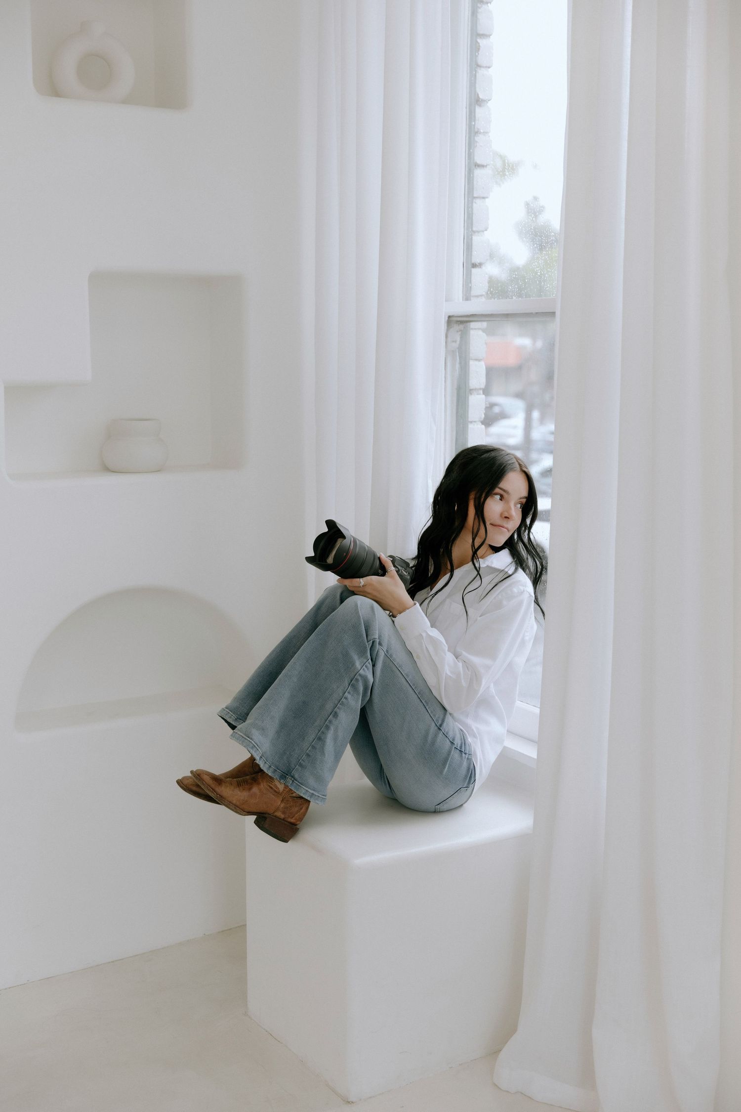 Photographer in white shirt and jeans sits casually on white windowsill holding camera in bright minimalist setting.