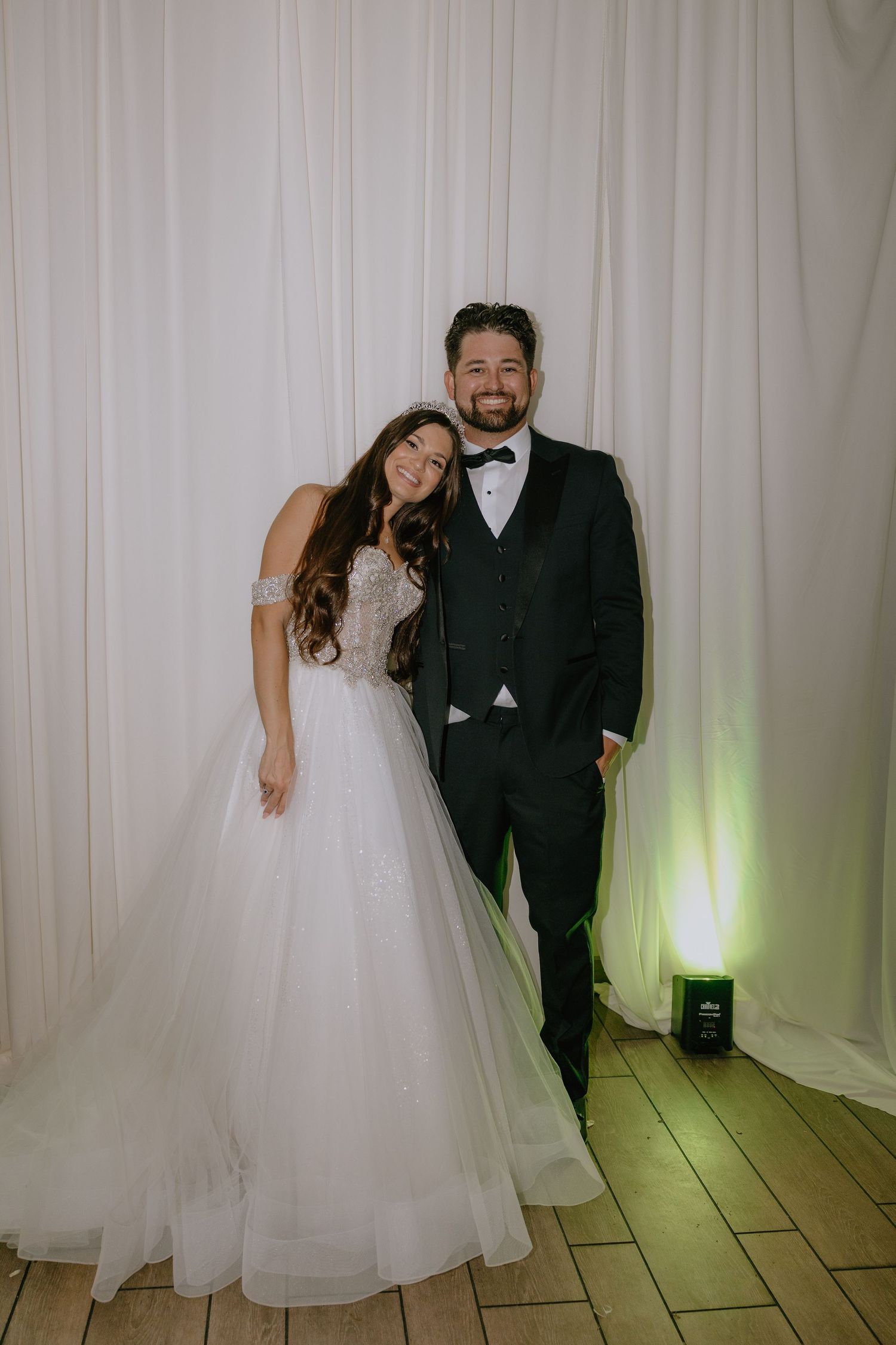 A smiling bride in a white ball gown and groom in a black tuxedo pose together in front of white drapes.
