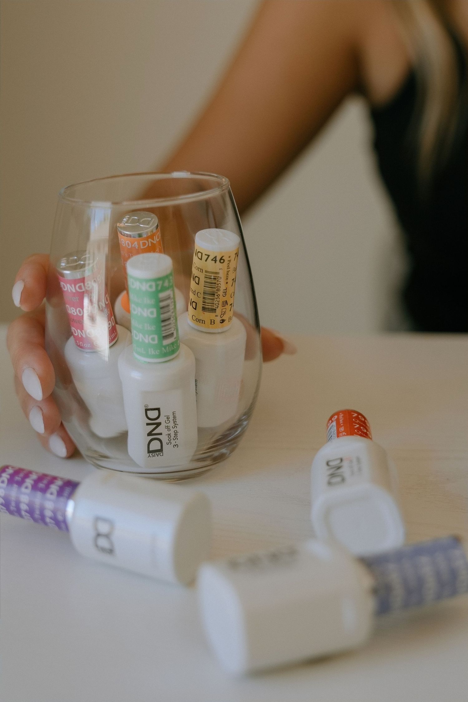 Woman holding a glass filled with DND gel nail polish bottles on a white table with more polishes nearby.